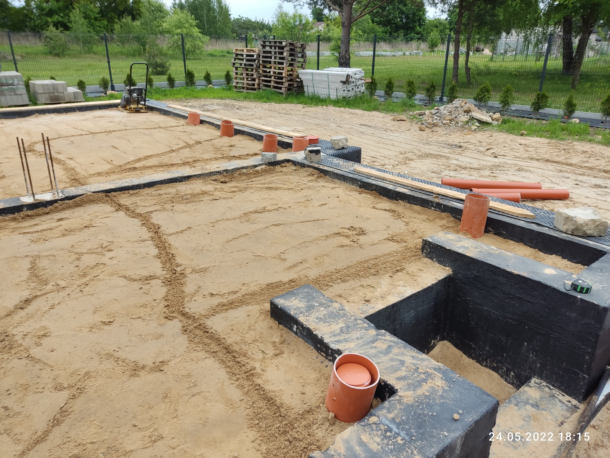 Przygotowanie fundamentów pod budynek: wykopane i zabezpieczone ściany fundamentowe, piasek wyrównany, widoczne rury kanalizacyjne, zagęszczarka spalinowa, palety i bloczki betonowe w tle.