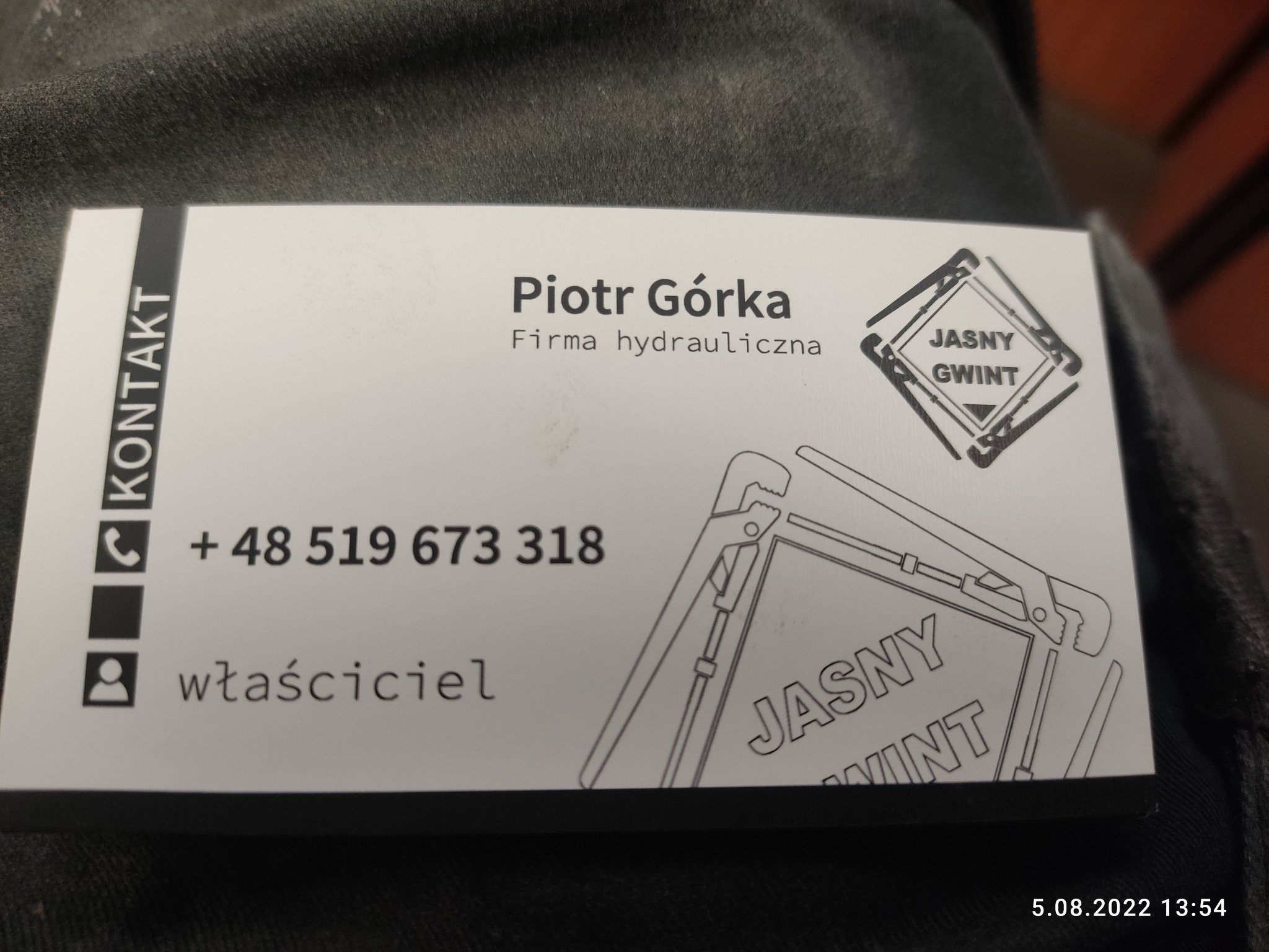Wizytówka firmy hydraulicznej 'Jasny Gwint' z danymi kontaktowymi Piotra Górki, właściciela, leżąca na ciemnym materiale.