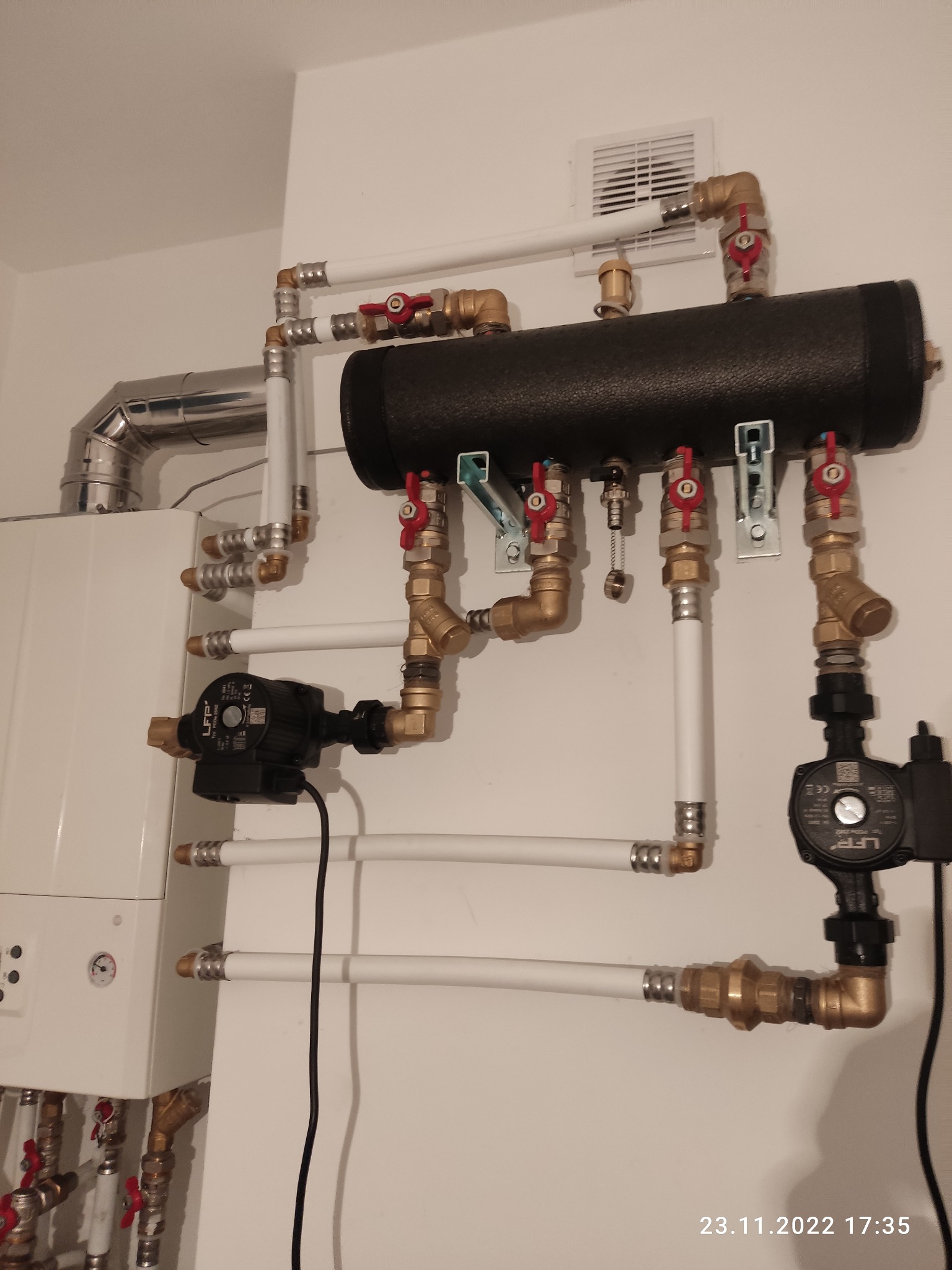 Rozbudowany system rur z zaworami i pompami, widoczny kocioł grzewczy, instalacja wewnątrz pomieszczenia z wentylacją.