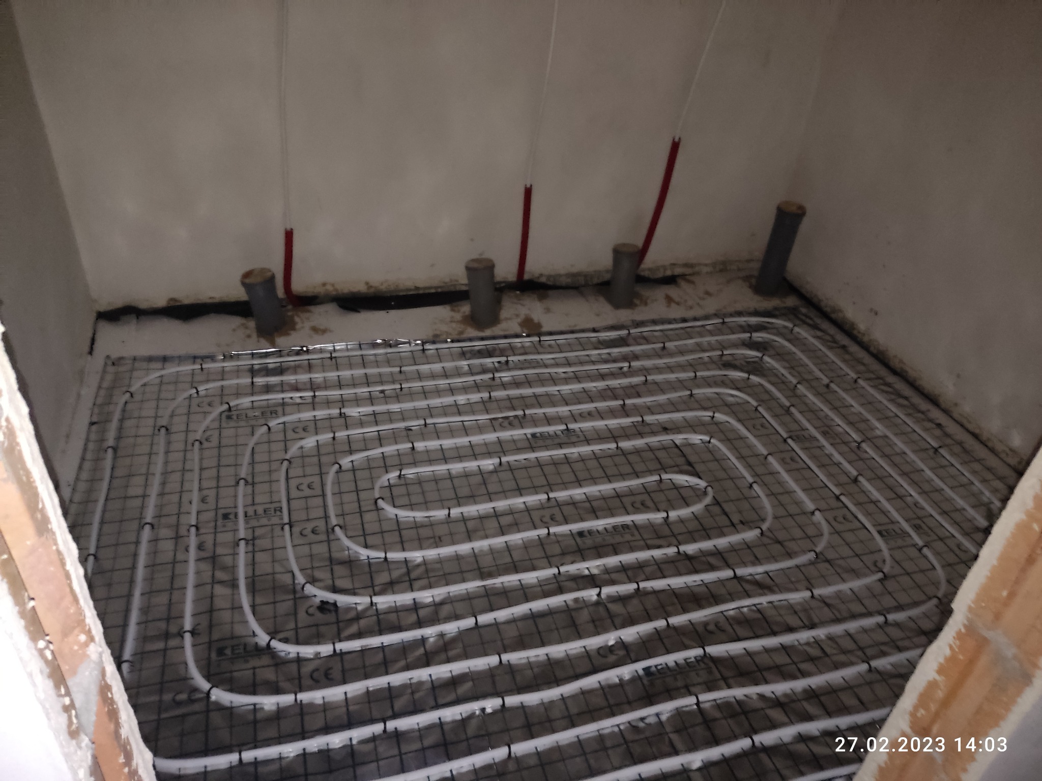 Rozłożona instalacja ogrzewania podłogowego na folii, rury marki Keller, widoczne pionowe rury przy ścianie, surowy stan wykończenia pomieszczenia.