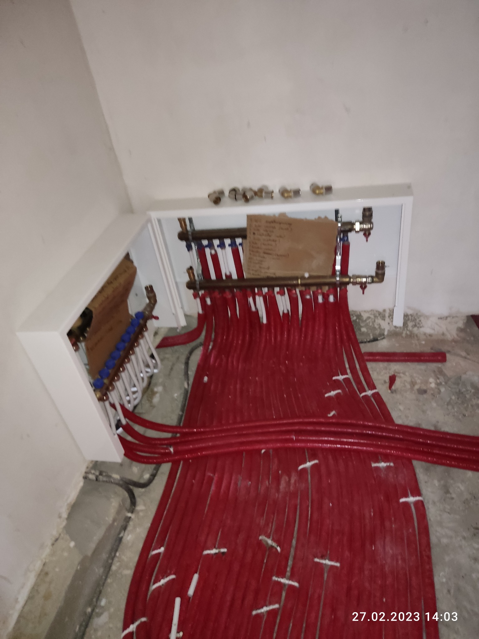 Rozdzielacz ogrzewania podłogowego z licznymi czerwonymi rurami ułożonymi na podłodze w surowym stanie, widoczne białe obudowy i mosiężne złączki, notatka z odręcznymi zapiskami na rozdzielaczu.