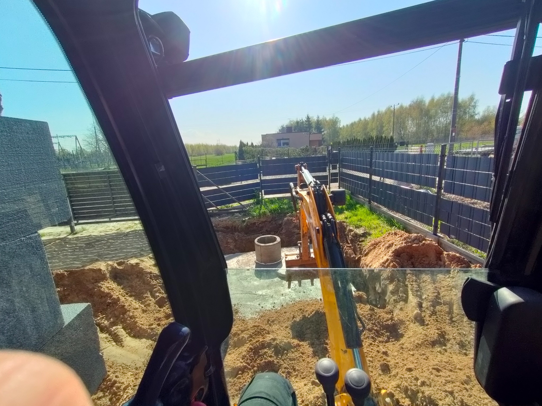 Widok z kabiny koparki na wykopany dół z betonowym kręgiem, w tle ogrodzenie panelowe i budynek.