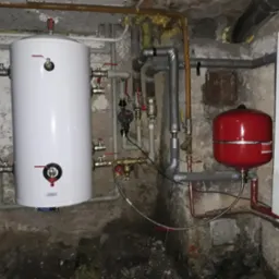 Instalacja grzewcza w piwnicy: biały zasobnik CWU, czerwone naczynie przeponowe i piec gazowy wiszący na tle surowych, betonowych ścian.