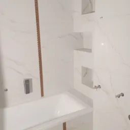 Biała łazienka z wanną obłożoną płytkami imitującymi marmur, z dekoracyjnymi listwami w kolorze miedzi i wnękami w ścianie.