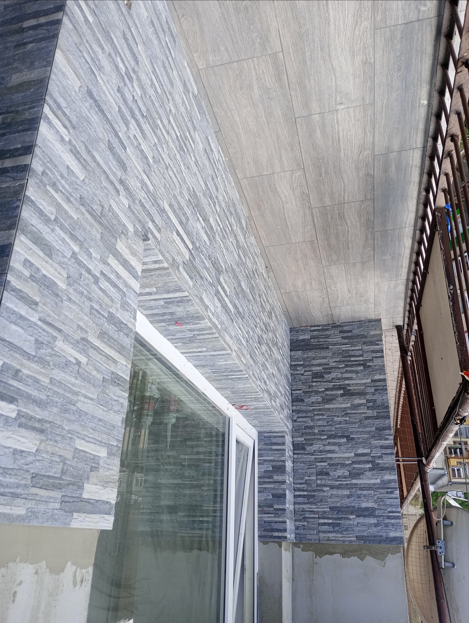 Wykończenie balkonu z szarą, kamienną okładziną ścian i sufitu imitującego drewno, widok z góry na balkon z białą ramą okienną.