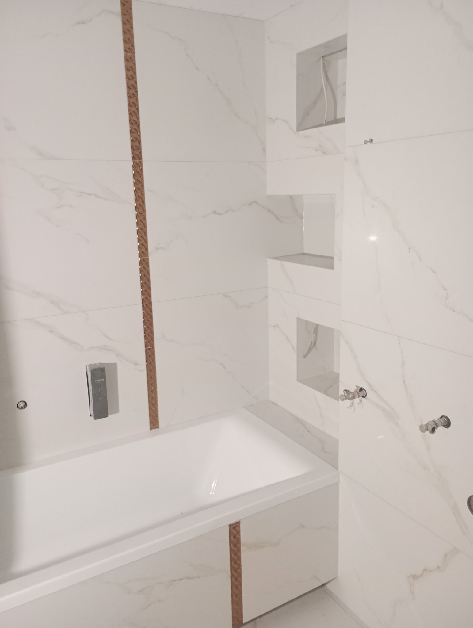 Biała łazienka z wanną obłożoną płytkami imitującymi marmur, z dekoracyjnymi listwami w kolorze miedzi i wnękami w ścianie.