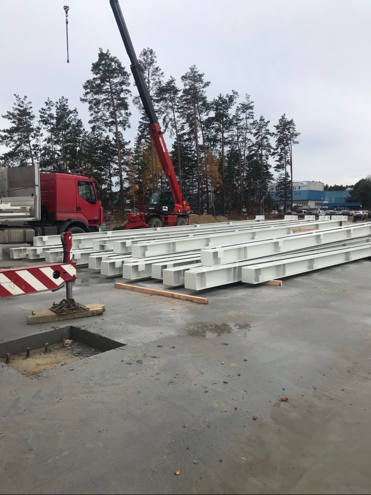 Przygotowane, białe, stalowe belki konstrukcyjne na placu budowy, widoczny czerwony dźwig teleskopowy i ciężarówka, w tle drzewa i budynek.