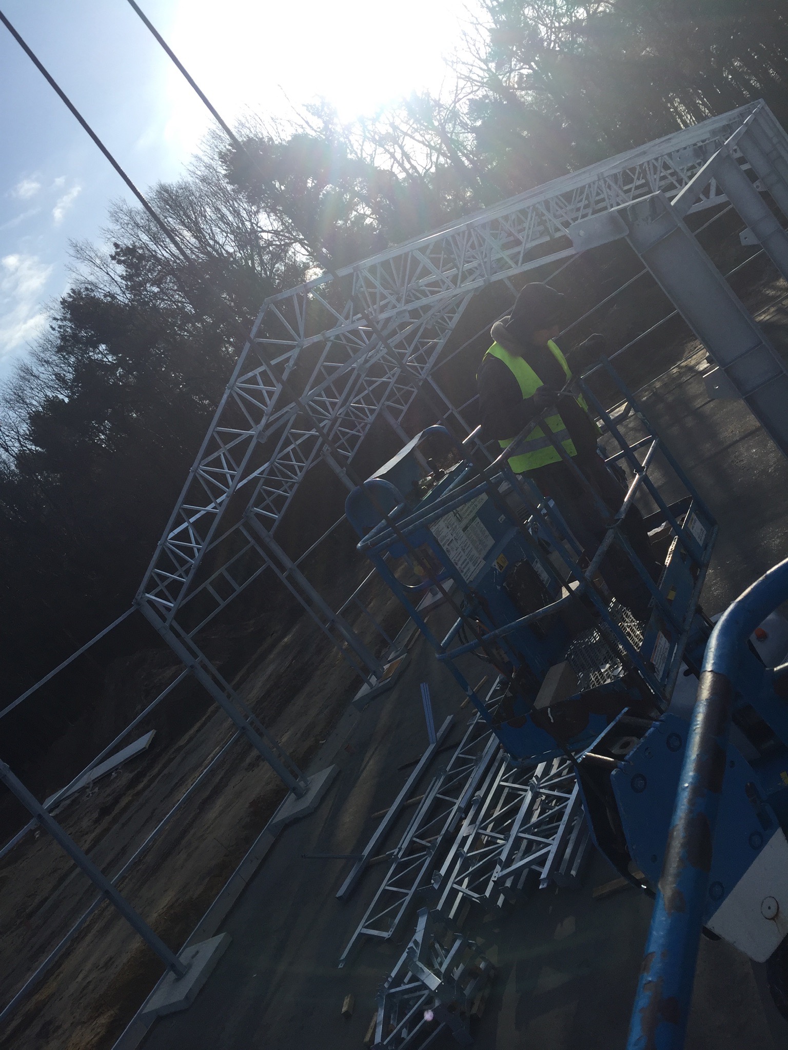 Pracownik w kamizelce odblaskowej na podnośniku koszowym montuje stalową konstrukcję hali na tle lasu i jasnego nieba.