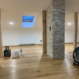 Wykończone wnętrze z drewnianą podłogą, kamienną kolumną i oknem dachowym, widoczne narzędzia i akcesoria malarskie.