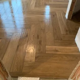 Ułożona w jodełkę, błyszcząca podłoga z płytek imitujących drewno, z fragmentem białych, sześciokątnych płytek mozaikowych przy progu i surowymi fragmentami ściany w tle.