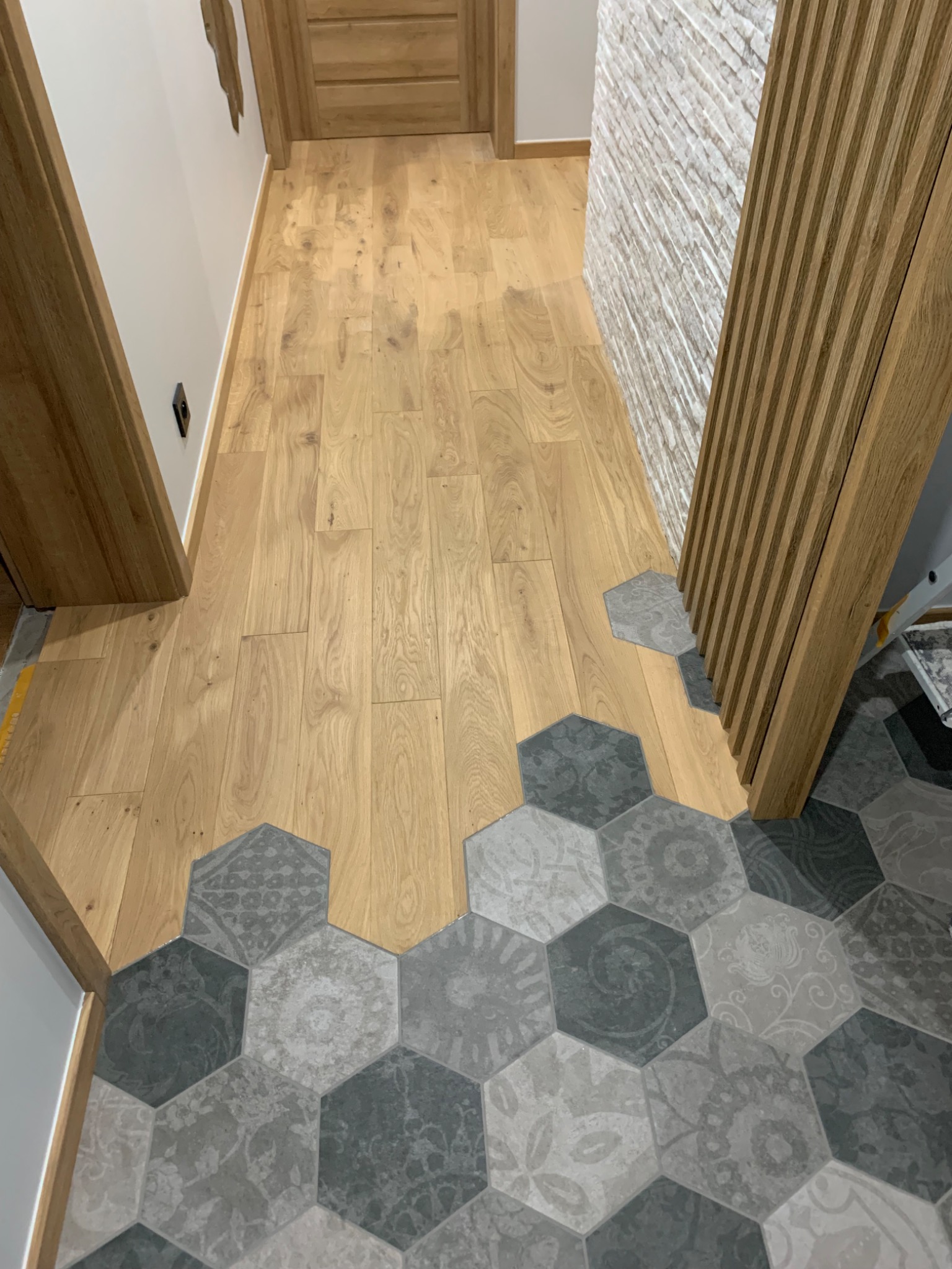 Połączenie drewnianej podłogi i heksagonalnych płytek w odcieniach szarości z dekoracyjnymi wzorami, widoczne drewniane listwy przypodłogowe i fragment ściany z dekoracyjną fakturą.