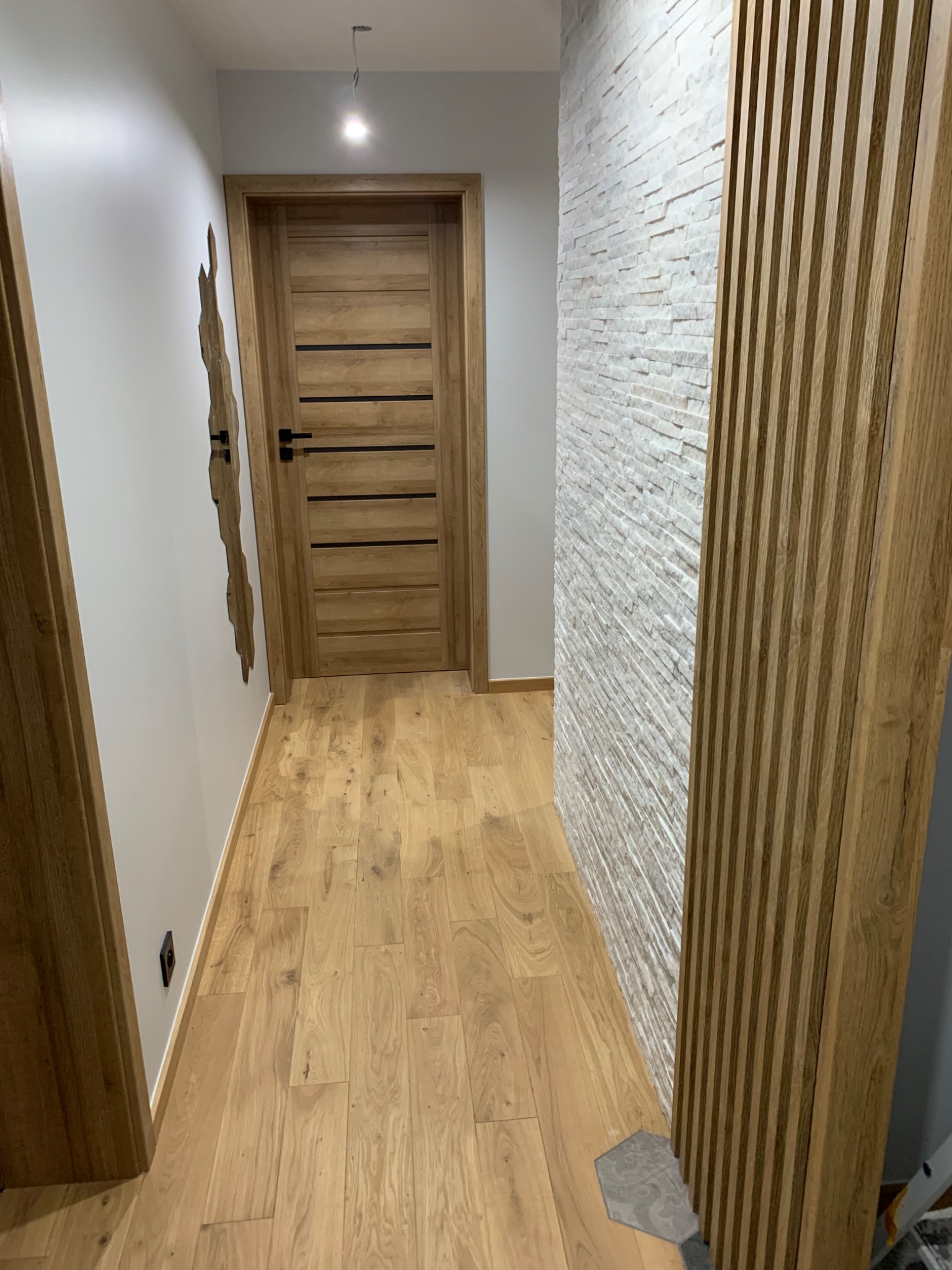 Wykończony korytarz z drewnianą podłogą, drzwiami z poziomymi wstawkami i ścianą z białej cegły oraz drewnianymi lamelami
