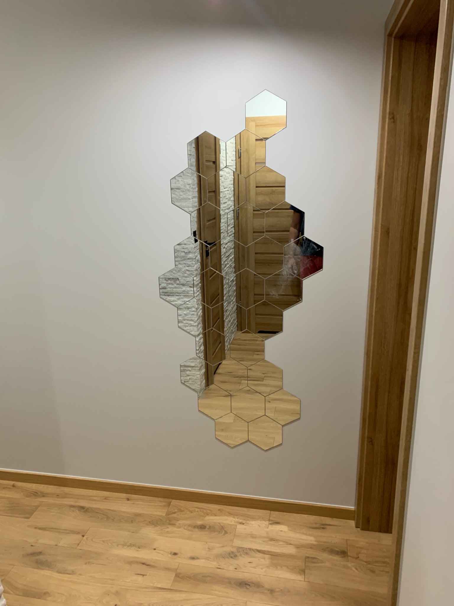 Mozaika lustrzana w kształcie plastra miodu na ścianie, odbijająca fragment wnętrza z drewnianymi elementami, w tle drewniana ościeżnica drzwiowa, na podłodze panele imitujące drewno.