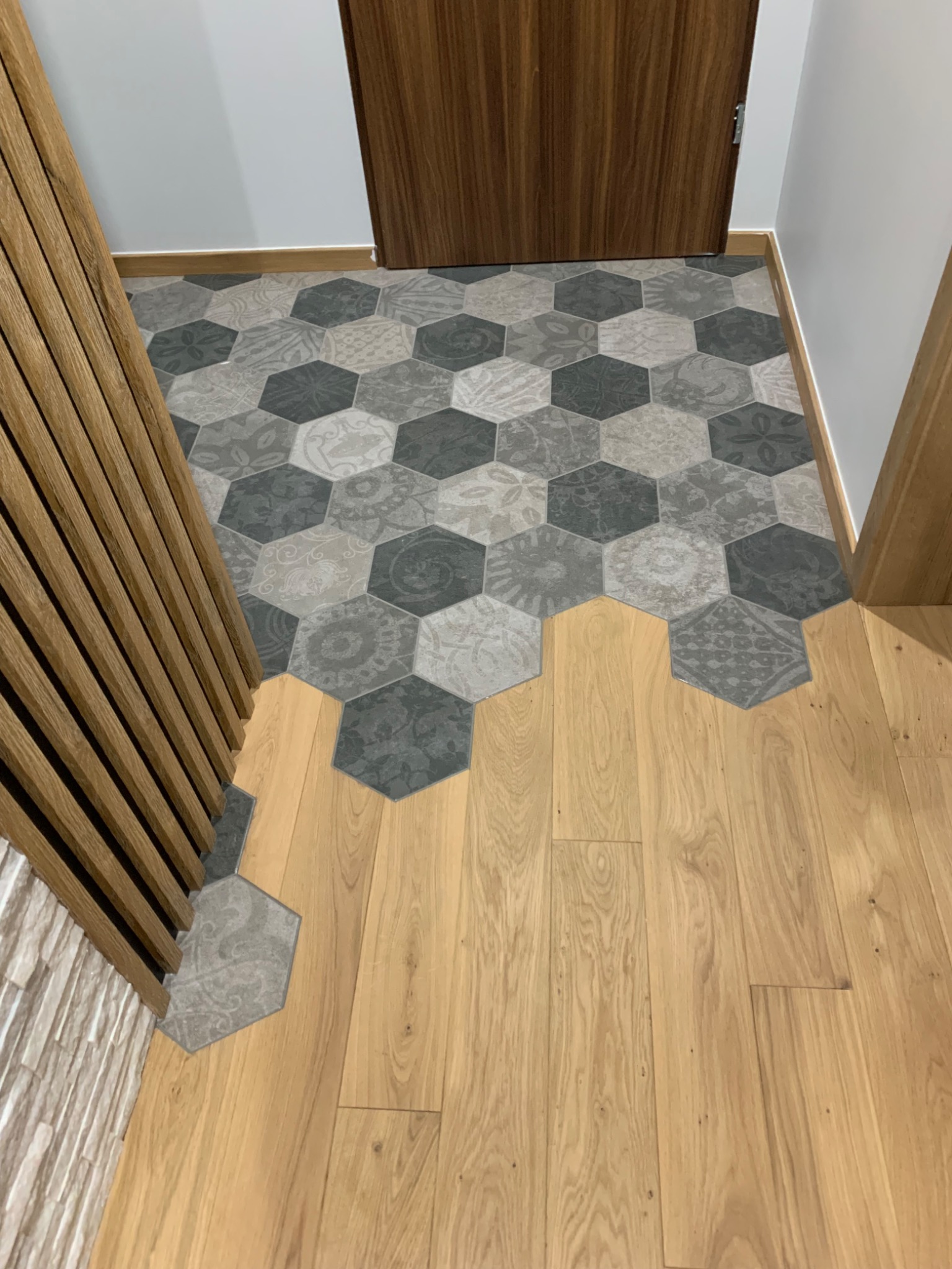 Połączenie heksagonalnych płytek ceramicznych w odcieniach szarości z wzorami i drewnianej podłogi w przedpokoju, widoczne drzwi z drewnianą okleiną i dekoracyjna drewniana listwa ścienna.