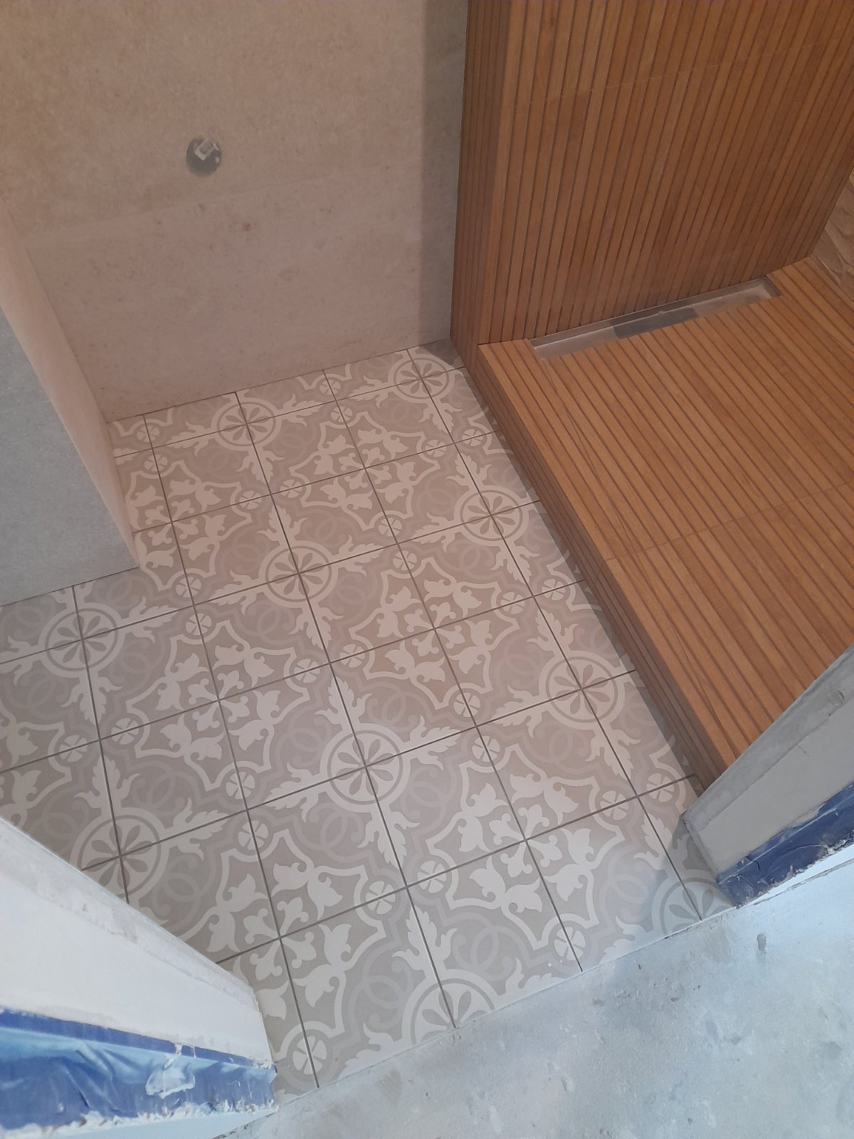 Ułożone płytki podłogowe w łazience z wzorem ornamentowym, obok drewniana zabudowa prysznica z odpływem liniowym.