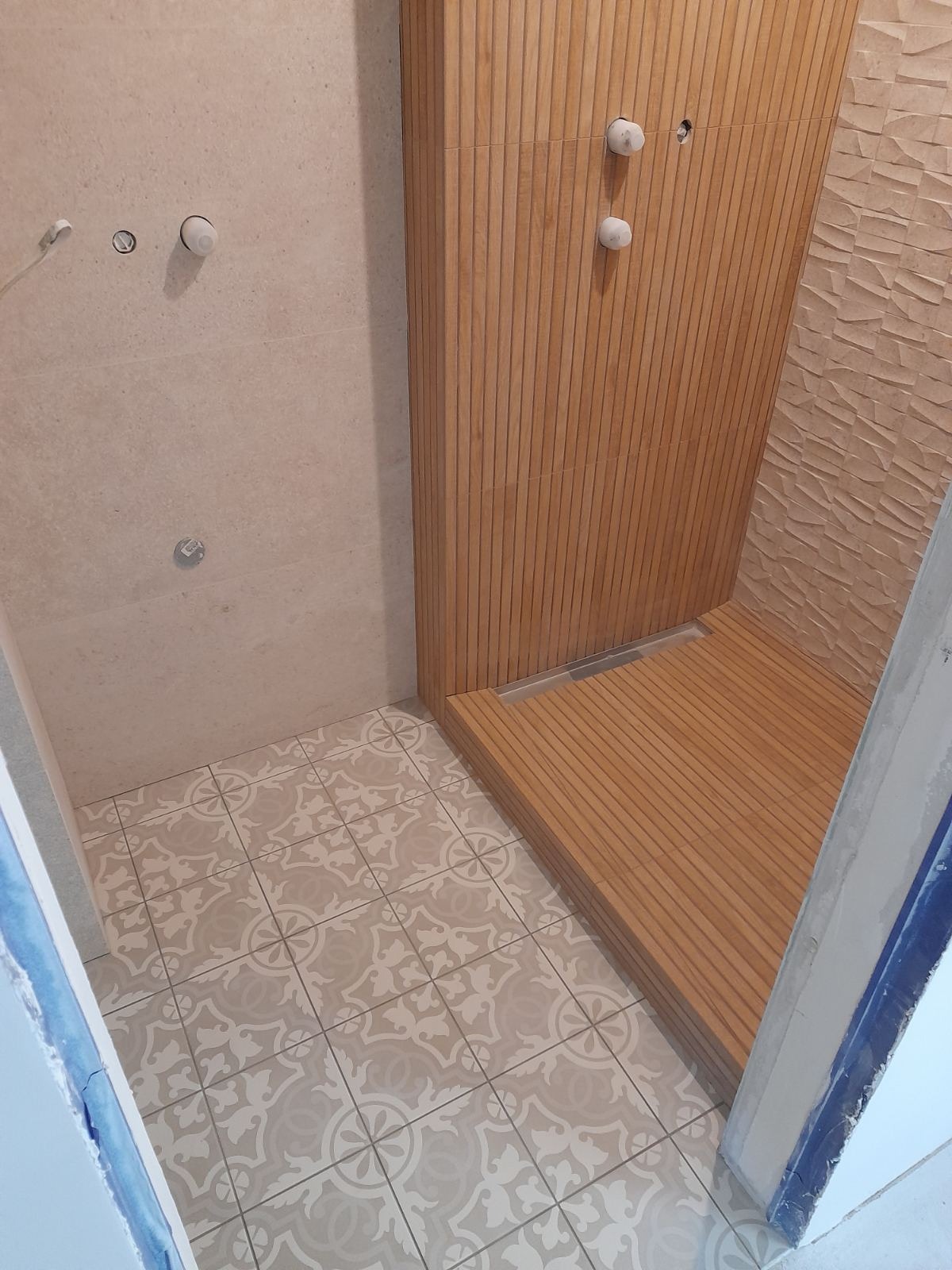 Wnętrze łazienki w trakcie prac wykończeniowych: drewniana obudowa prysznica z odpływem liniowym, beżowe płytki ścienne z wypukłą fakturą, podłoga wyłożona dekoracyjnymi płytkami w odcieniach...