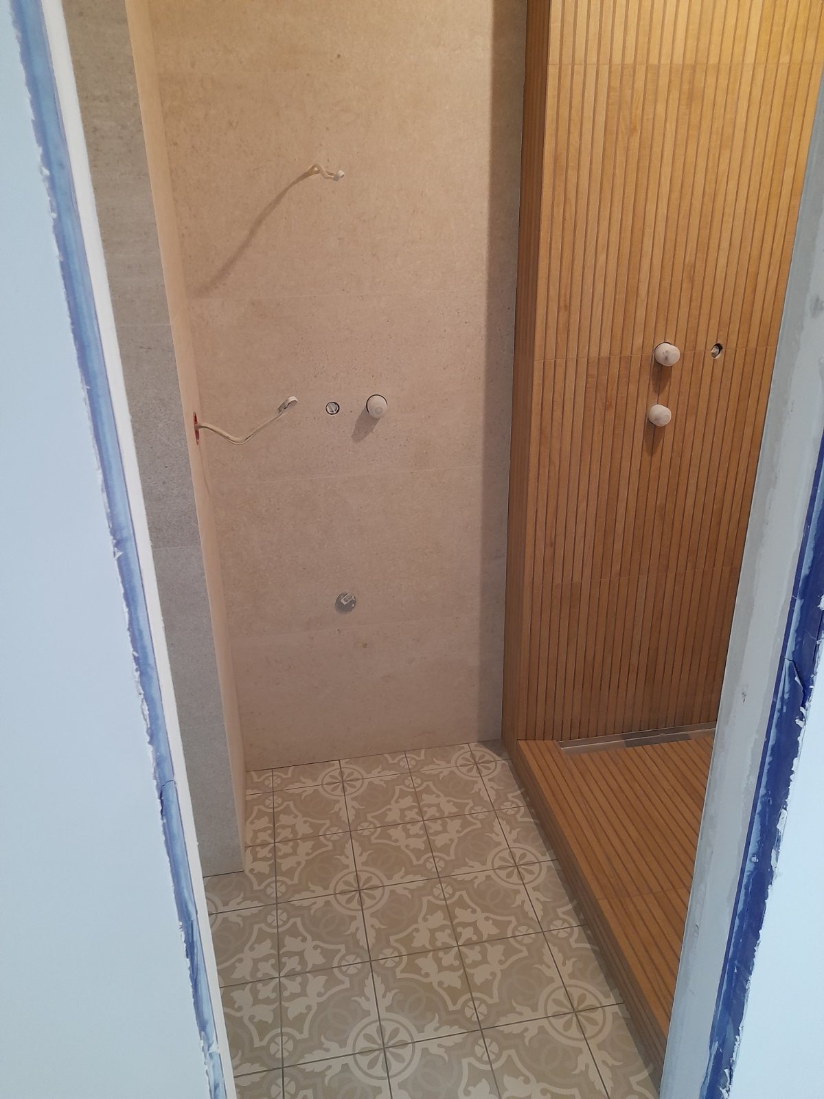 Niedokończona łazienka z beżowymi płytkami na ścianach, wzorzystą podłogą i drewnianą kabiną prysznicową z odpływem liniowym; widoczne wystające kable i rury instalacyjne.
