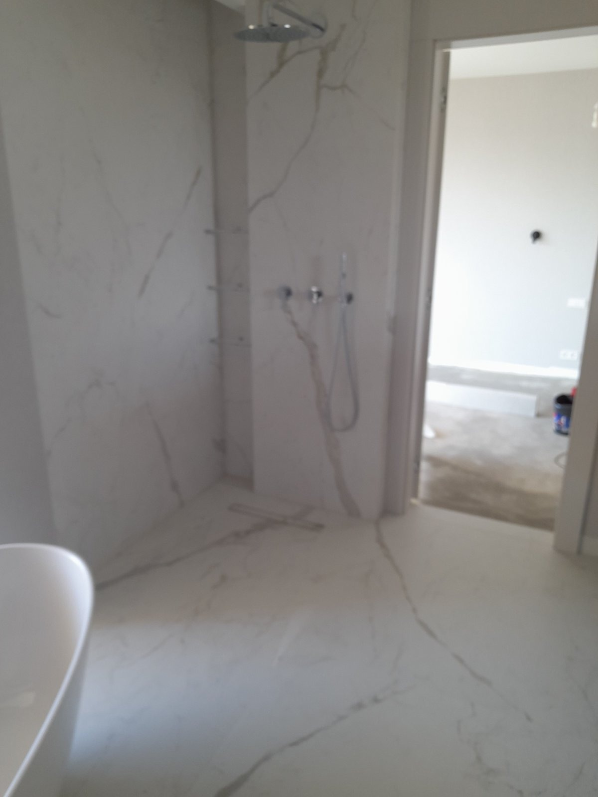 Nowoczesna łazienka w trakcie wykańczania z marmurowymi płytkami, widoczna kabina prysznicowa typu walk-in z deszczownicą, częściowo widoczna wanna wolnostojąca i otwarte drzwi do kolejnego...