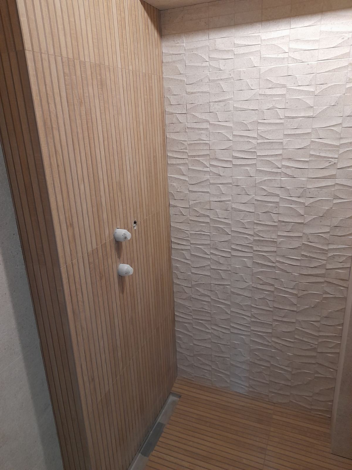 Wykończona kabina prysznicowa z drewnopodobnymi panelami ściennymi z pionowymi żłobieniami, kontrastującymi z trójwymiarową, jasną płytką na sąsiedniej ścianie oraz drewnianą podłogą, widoczne...