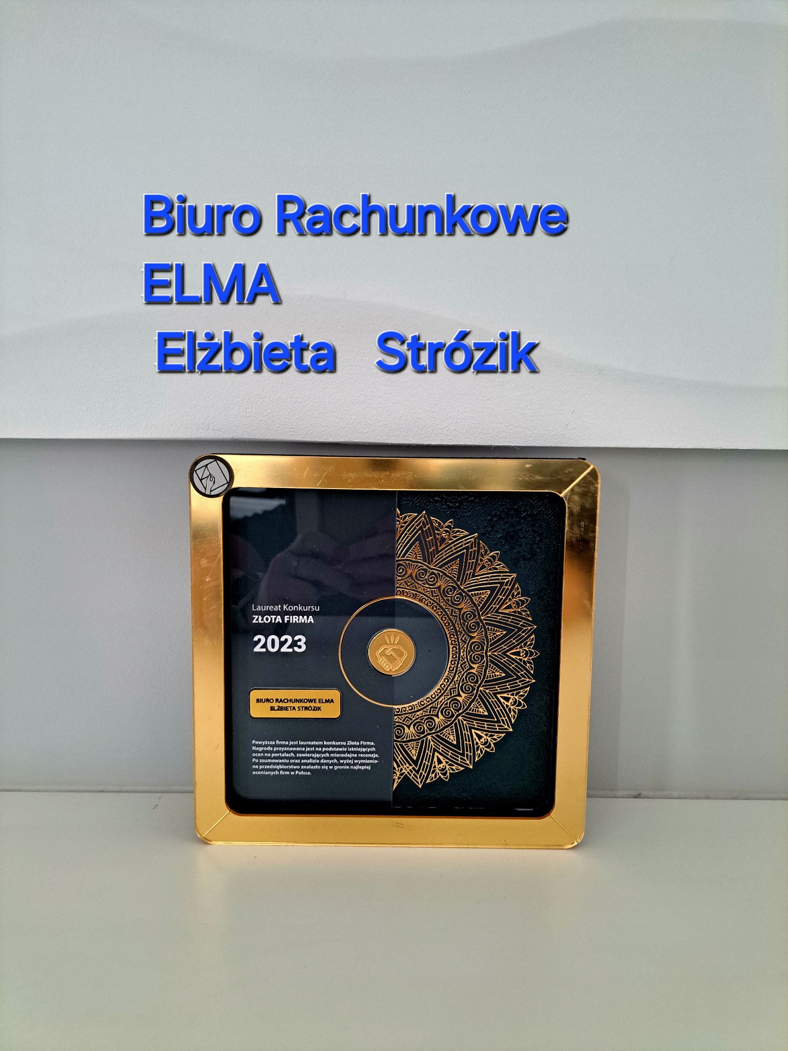 Tablica z informacją o nagrodzie 'Złota Firma 2023' dla Biura Rachunkowego ELMA Elżbieta Strózik, umieszczona na jasnej ścianie.