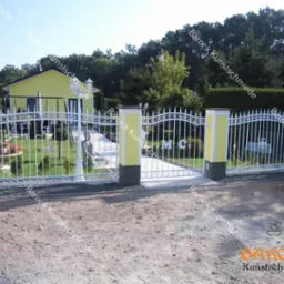 Biała, ozdobna brama wjazdowa z żółtymi słupkami i monogramem 'MC', prowadząca do zadbanego ogrodu z żółtym domem w tle.