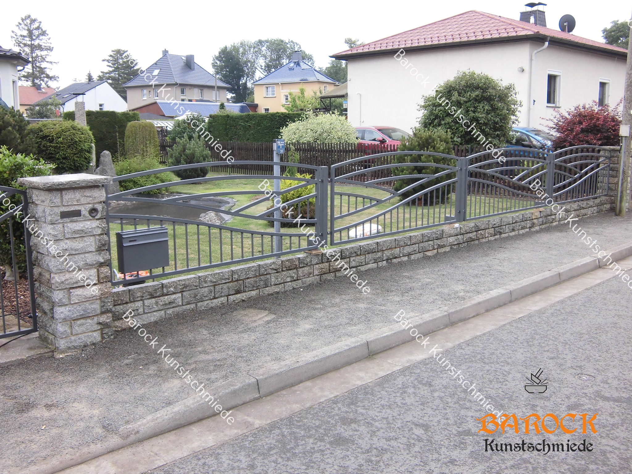 Szara, metalowa konstrukcja ogrodzenia z dekoracyjnymi elementami, osadzona na niskim murku z kamienia łupanego, z widoczną skrzynką na listy, na tle domów jednorodzinnych i zieleni.