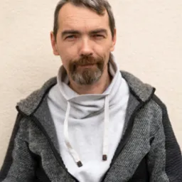 Portret mężczyzny z brodą i wąsami, ubrany w szarą bluzę z kapturem i rozpinaną kurtkę o fakturze warkocza na tle beżowej ściany.