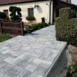 Ułożona szara kostka brukowa prowadząca do domu z brązowym dachem, widoczny drewniany płotek i ozdobne krzewy.