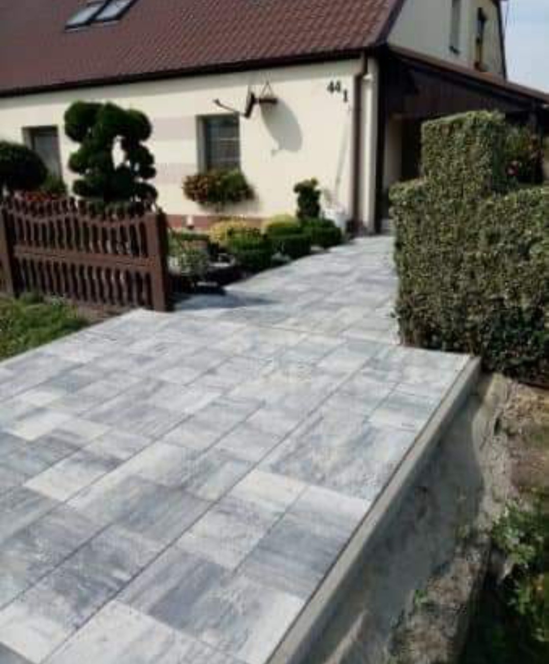 Ułożona szara kostka brukowa prowadząca do domu z brązowym dachem, widoczny drewniany płotek i ozdobne krzewy.