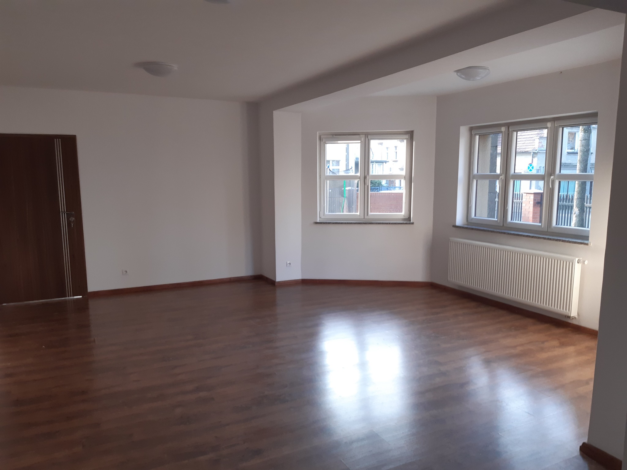 Pusty pokój po remoncie z parkietem, białymi ścianami, dwoma oknami z grzejnikiem i brązowymi drzwiami.