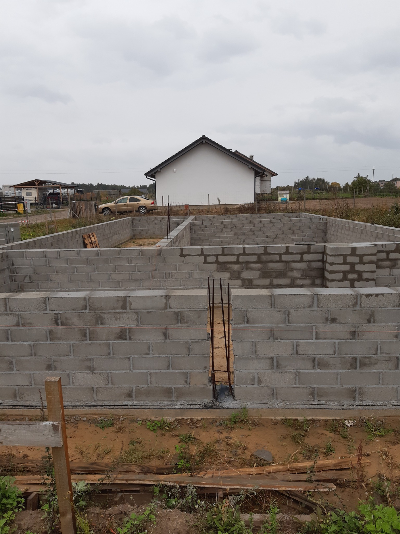 Wykopane fundamenty z bloczków betonowych, widoczne pręty zbrojeniowe, w tle dom w stanie surowym zamkniętym i samochód osobowy