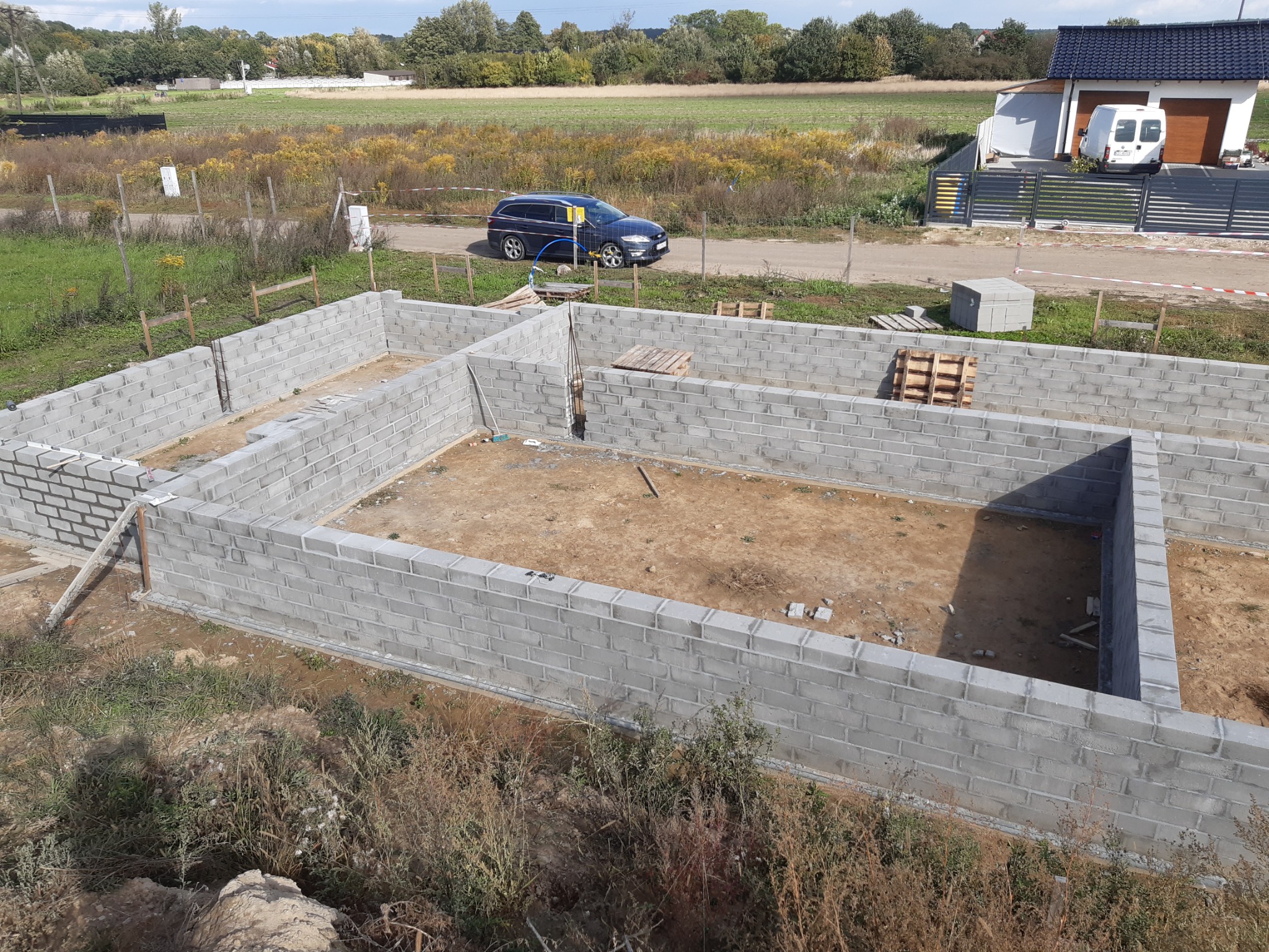Fundamenty domu jednorodzinnego w trakcie budowy, widok z góry na szare bloczki betonowe, w tle krajobraz wiejski z samochodem osobowym i budynkiem gospodarczym.
