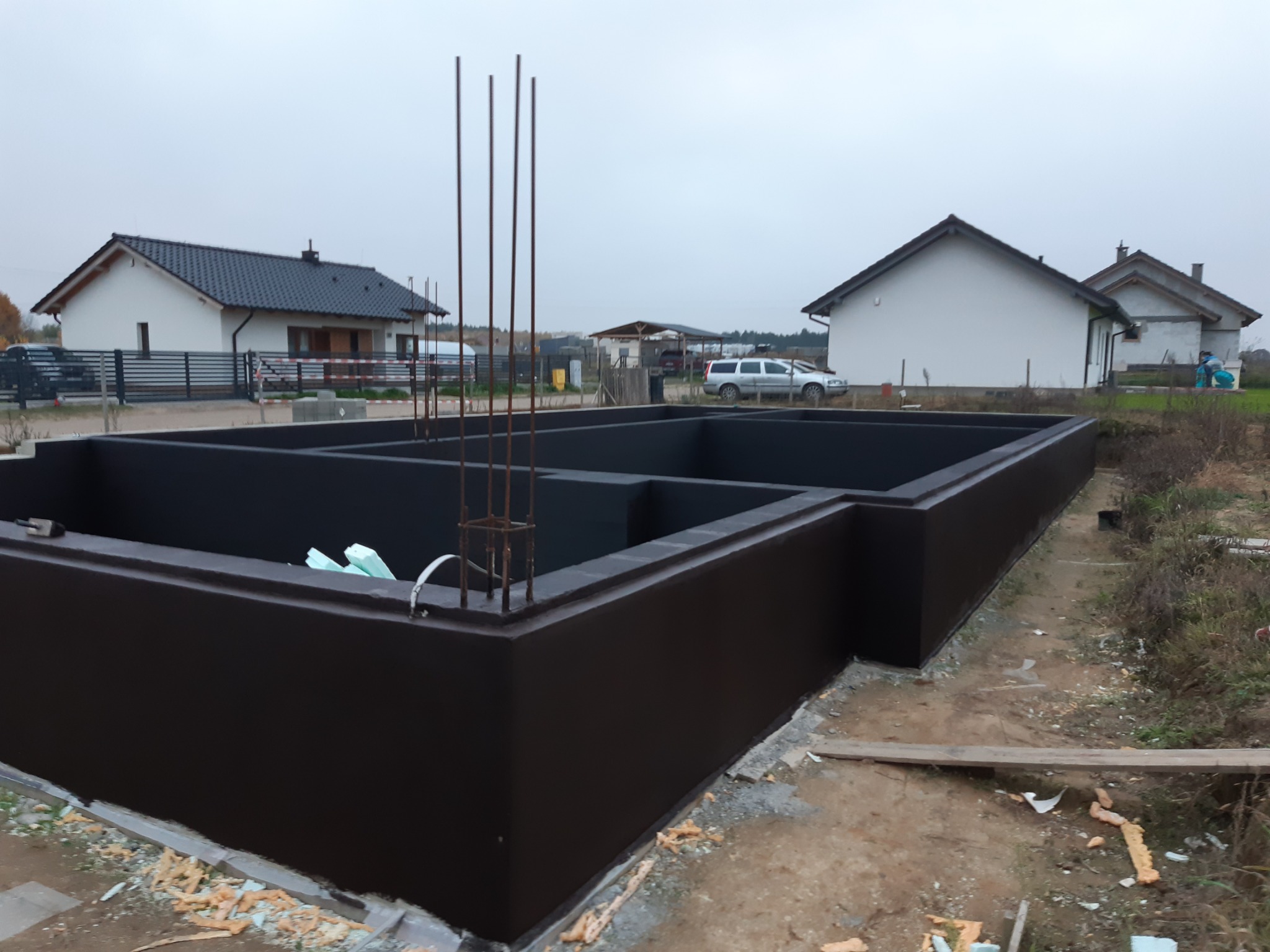 Zabezpieczone fundamenty domu jednorodzinnego z wystającymi prętami zbrojeniowymi, widoczne fragmenty izolacji termicznej i przygotowany teren pod budowę.