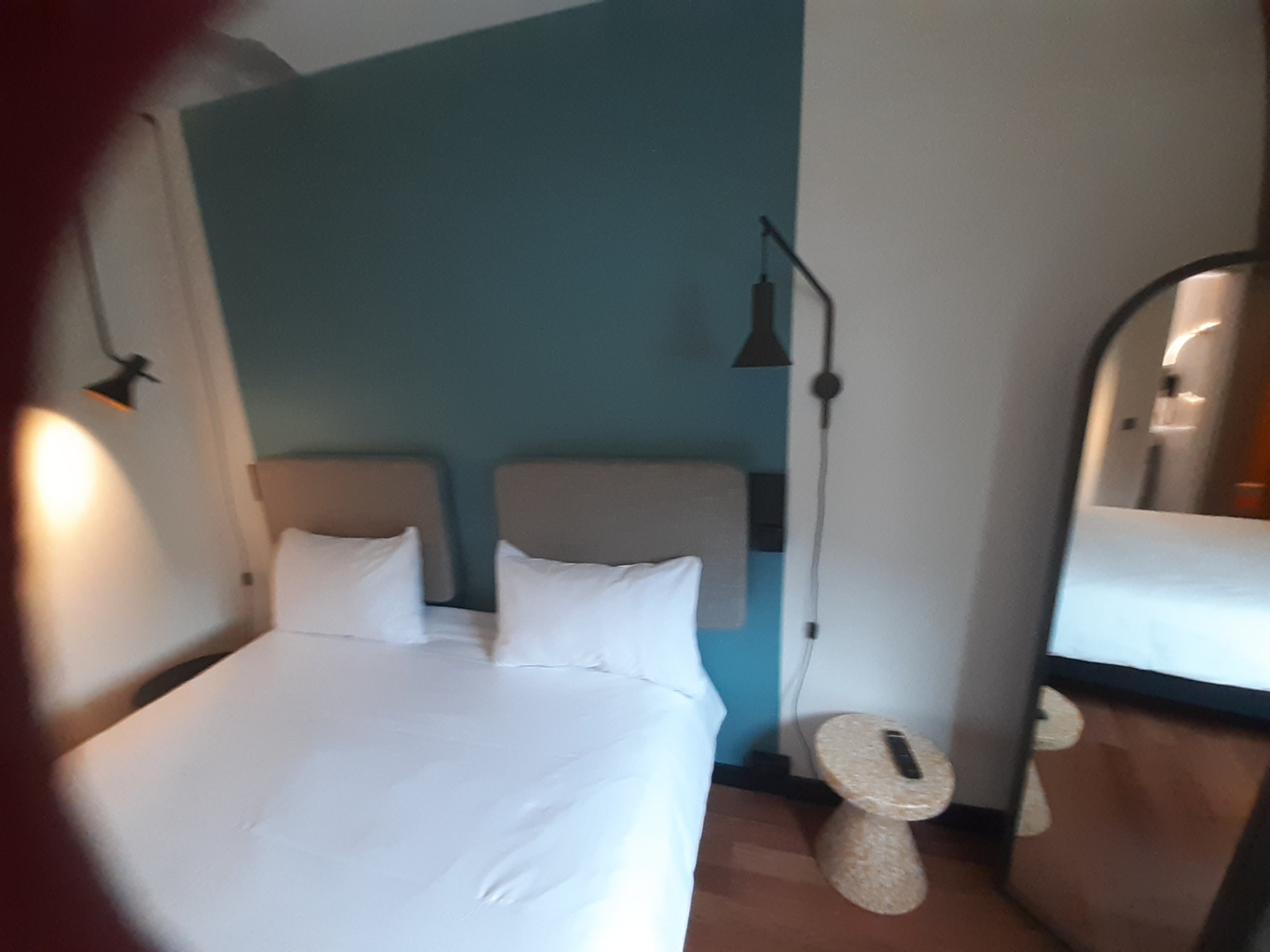 Nowoczesna sypialnia po remoncie: łóżko z białą pościelą na tle turkusowej ściany, lampka nocna z czarnym kloszem, stolik z pilotem, duże lustro odbijające wnętrze pokoju.