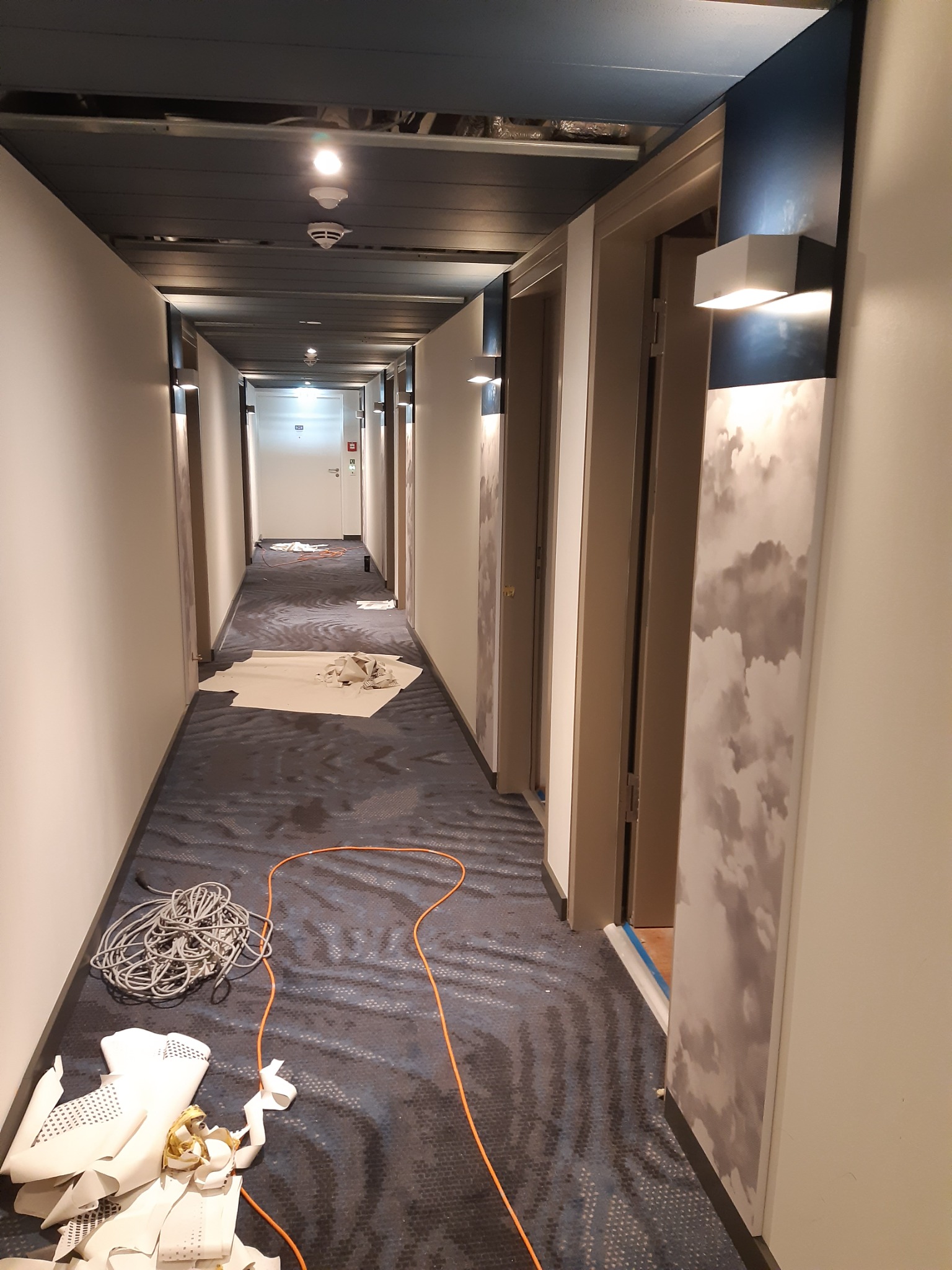Korytarz w trakcie prac wykończeniowych: widoczne przewody elektryczne, resztki materiałów budowlanych na podłodze, częściowo zamontowane oświetlenie ścienne i panele dekoracyjne z motywem chmur.