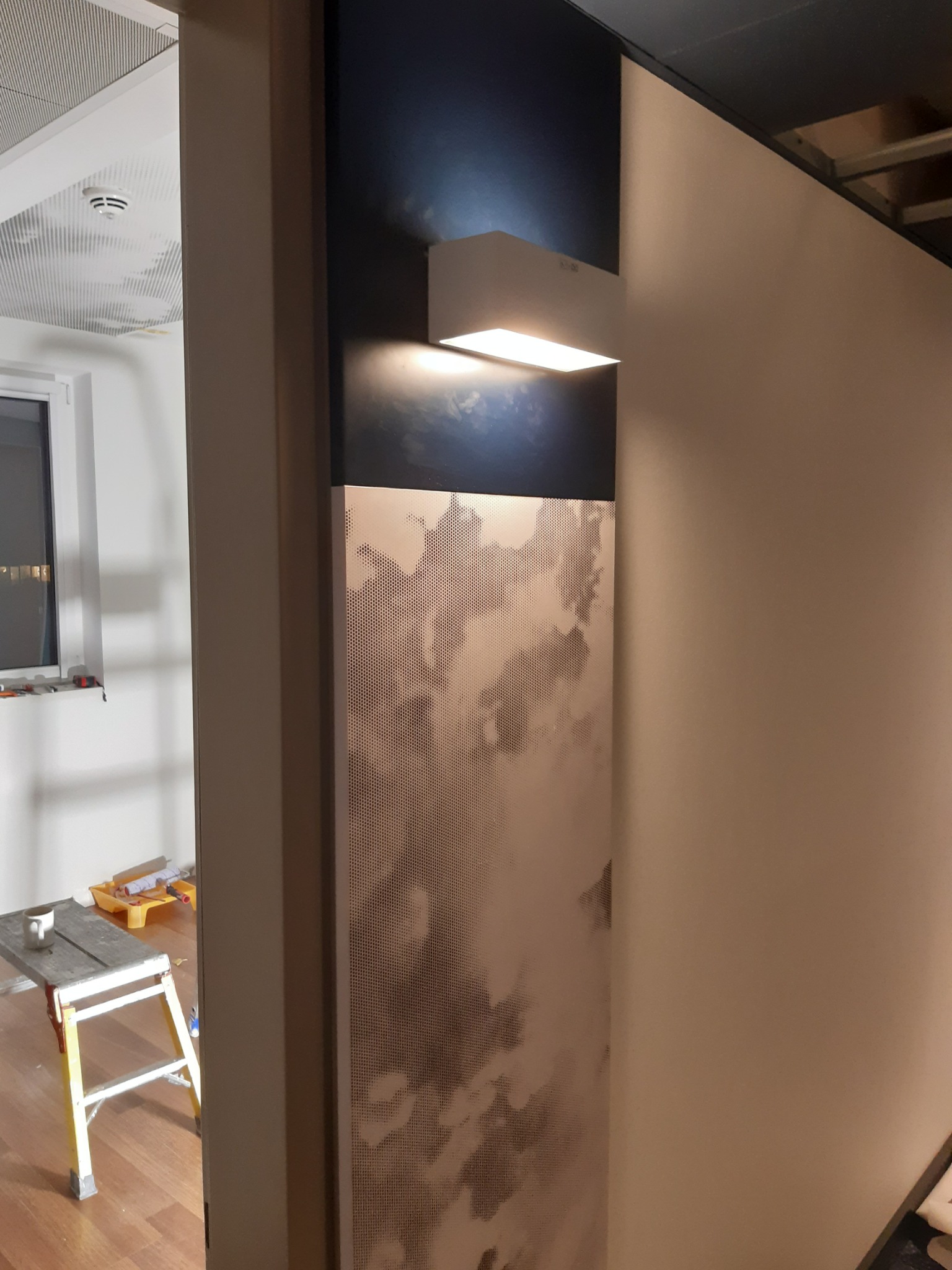Fragment ściany z zamontowanym panelem dekoracyjnym z perforacją i podświetleniem LED, widoczny drabinka malarska w tle.