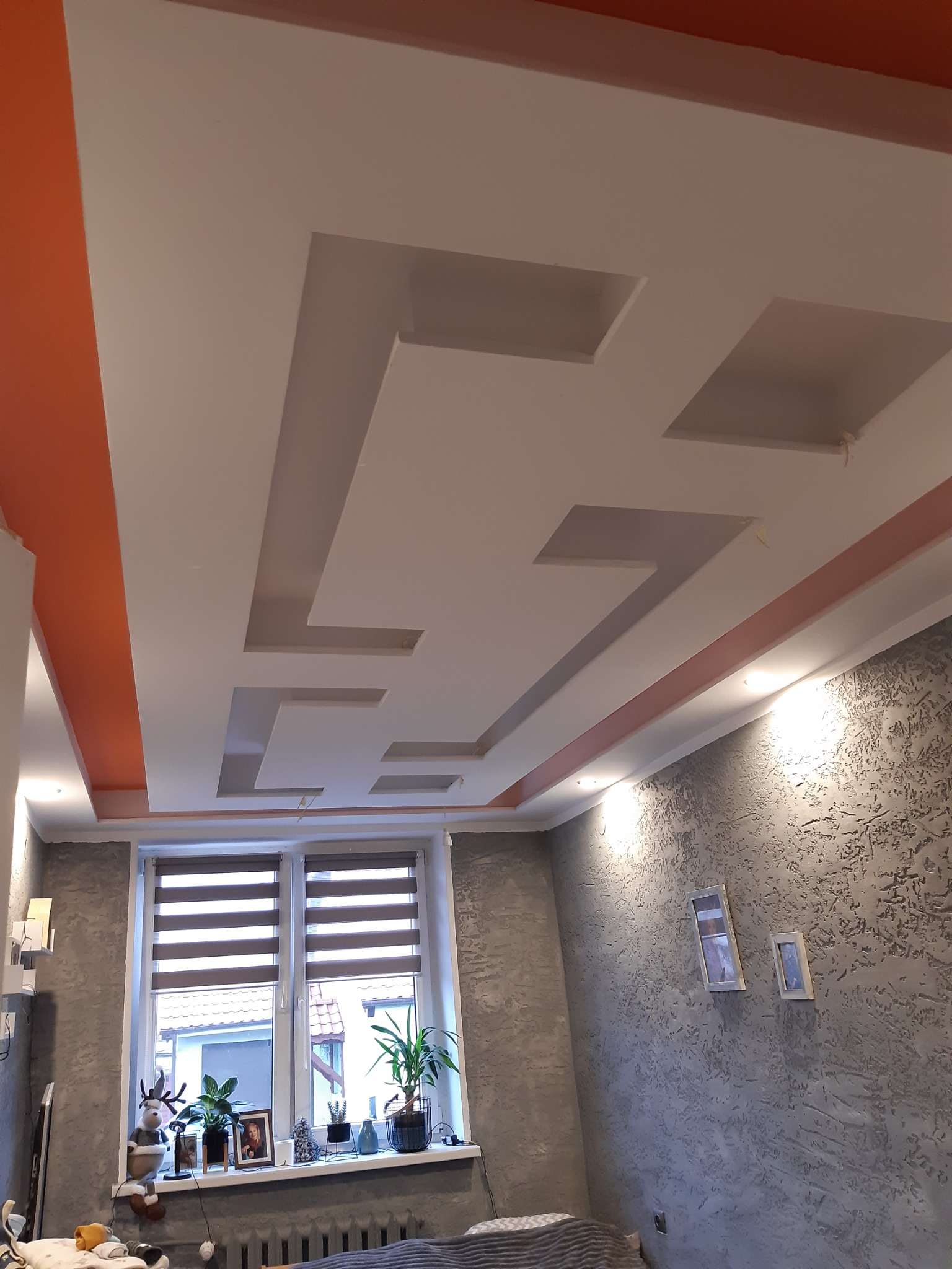 Nowoczesny sufit podwieszany z geometrycznymi wgłębieniami i pomarańczowymi akcentami, widok z dołu na szarą ścianę z dekoracyjną fakturą i oknem z roletami.
