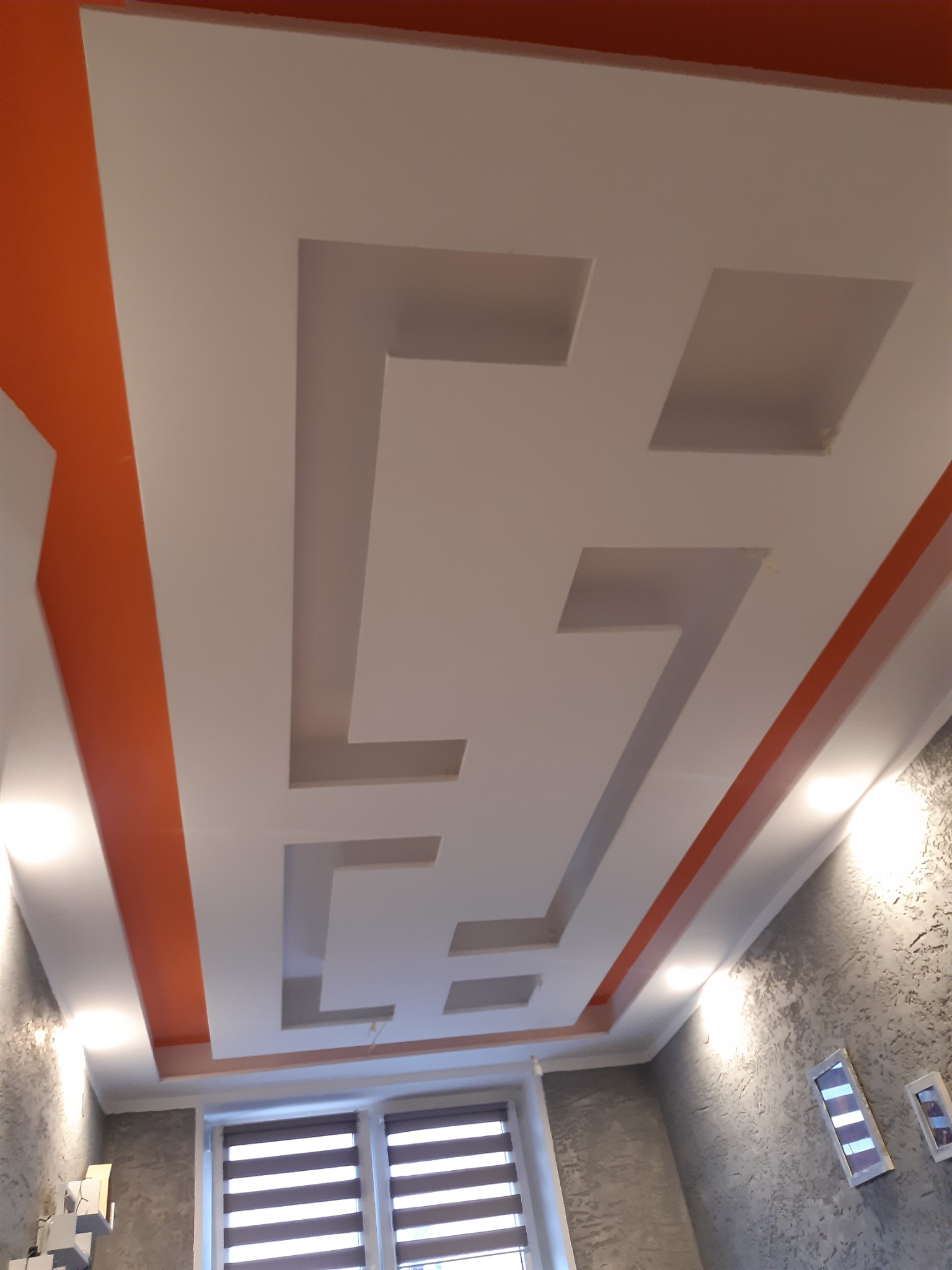 Dekoracyjny sufit podwieszany z geometrycznym wzorem, pomalowany na biało z pomarańczowymi akcentami, widok z dołu na okno z żaluzjami.