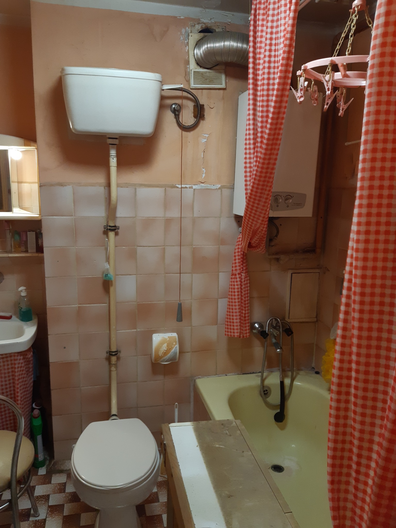 Wnętrze łazienki z toaletą, wanną i bojlerem, widoczne ślady użytkowania i niedoskonałości wykończenia, zasłonka w czerwoną kratę.