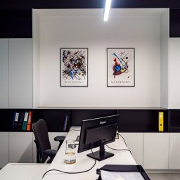 Kancelaria Radców Prawnych Tomasz Czapczyński Sp. k. - Nowoczesne biuro z białymi szafkami, czarnymi półkami, dwoma abstrakcyjnymi obrazami Kandinsky'ego na ścianie, biurkiem z monitorem Iiyama, szklanką wody i segregatorami w półkach.