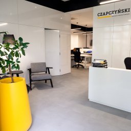 Kancelaria Radców Prawnych Tomasz Czapczyński Sp. k. - Nowoczesne wnętrze kancelarii prawnej z recepcją, szarym fotelem i dużą żółtą donicą z rośliną, widok na biura w tle.