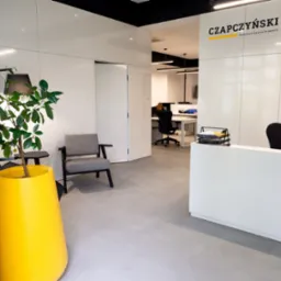Nowoczesne wnętrze kancelarii prawnej z recepcją, szarym fotelem i dużą żółtą donicą z rośliną, widok na biura w tle.