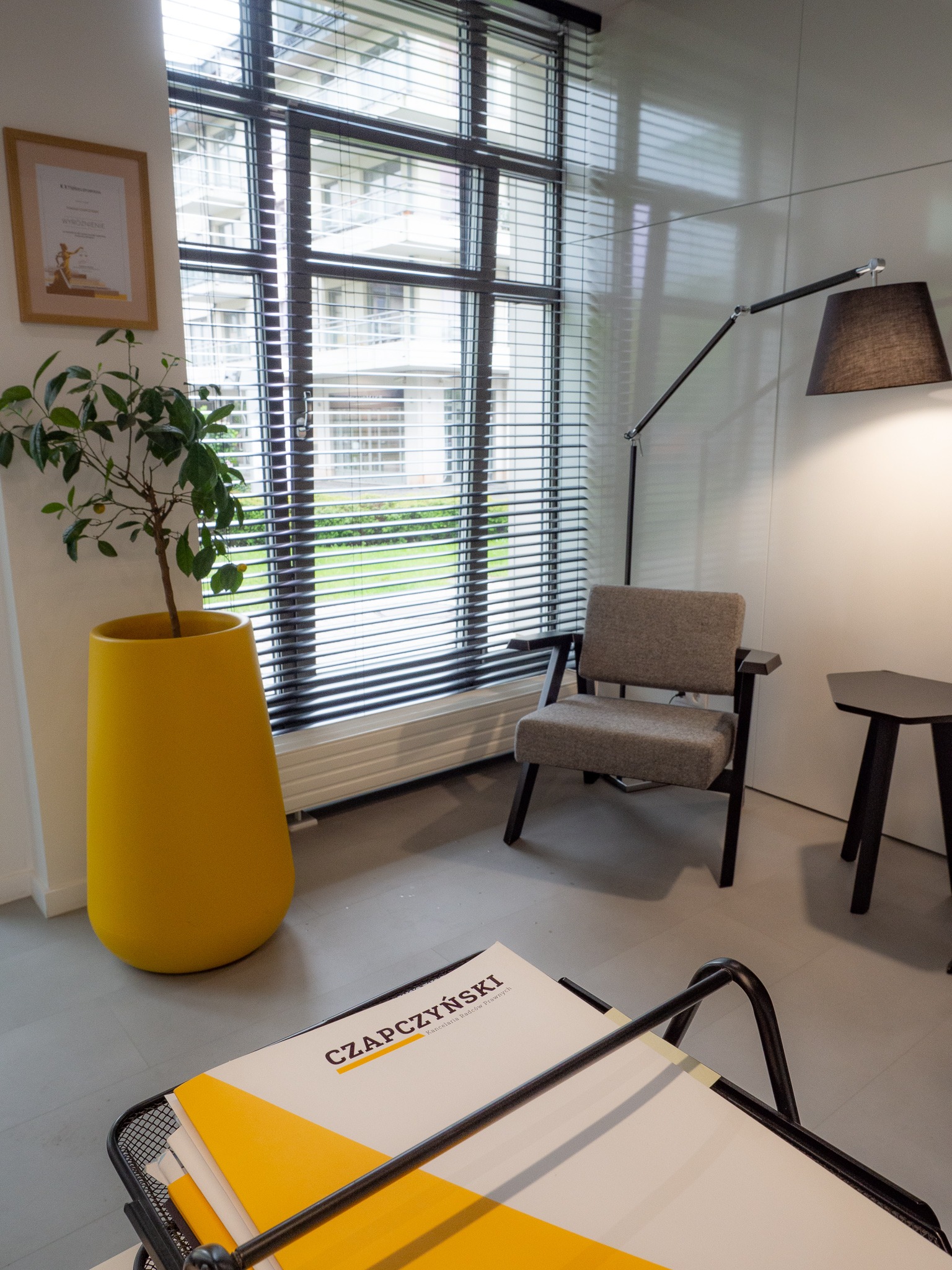 Wnętrze biura: Szare krzesło na tle okna z żaluzjami, obok lampa podłogowa i stolik, w rogu żółta donica z drzewkiem cytrynowym, na pierwszym planie dokumenty z nazwą 'CZAPCZYŃSKI Kancelaria Radców...