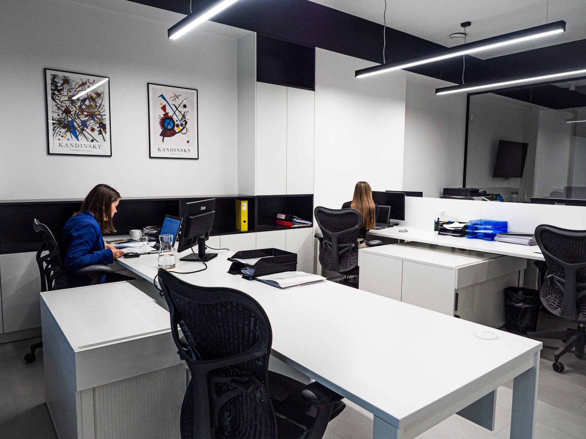 Nowoczesne biuro z pracownikami przy biurkach, minimalistyczny wystrój, obrazy Kandinsky'ego na ścianie, oświetlenie ledowe.