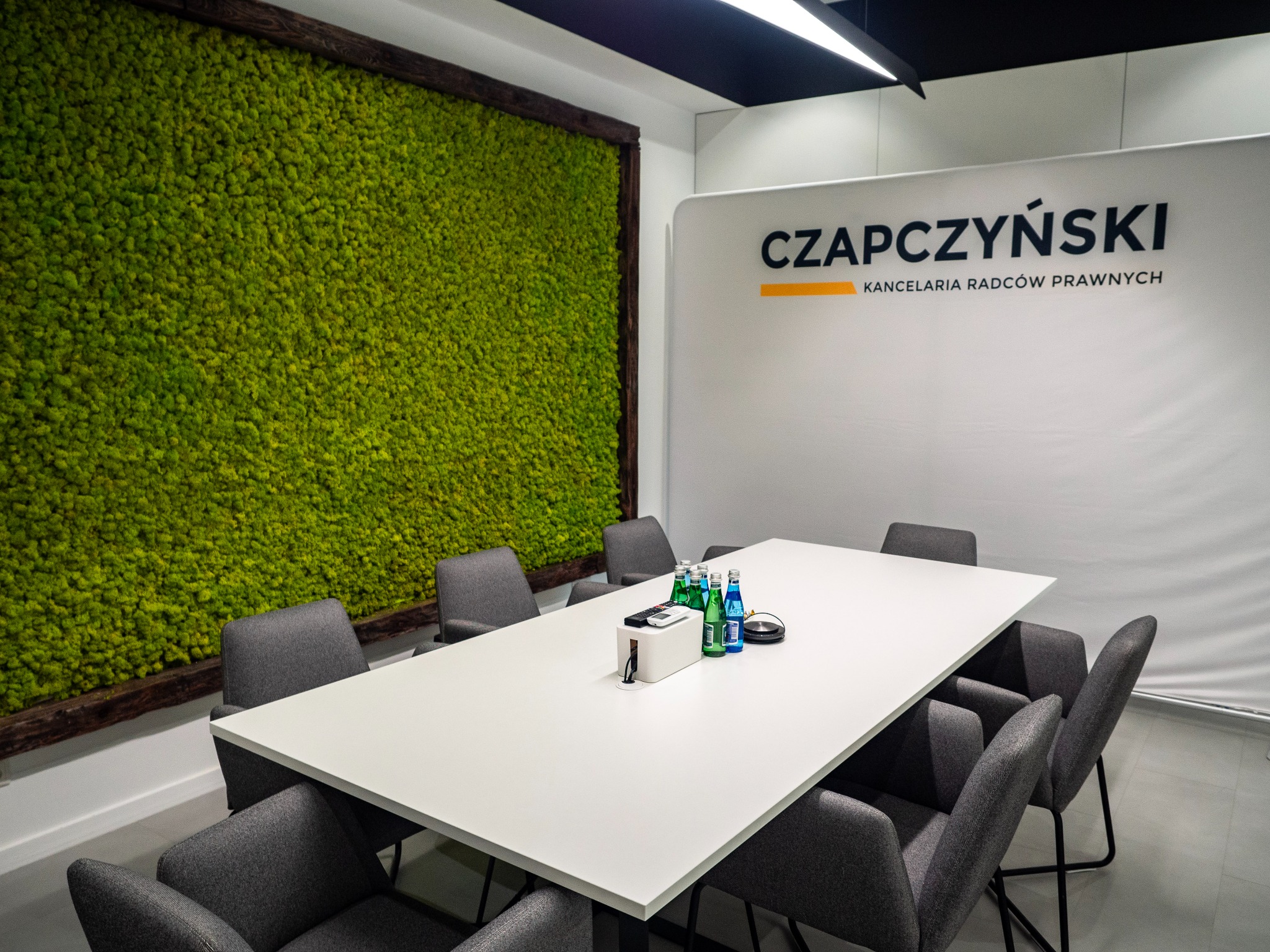 Sala konferencyjna z długim białym stołem otoczonym szarymi krzesłami, ściana z mchu w drewnianej ramie i logo 'Czapczyński Kancelaria Radców Prawnych' w tle.