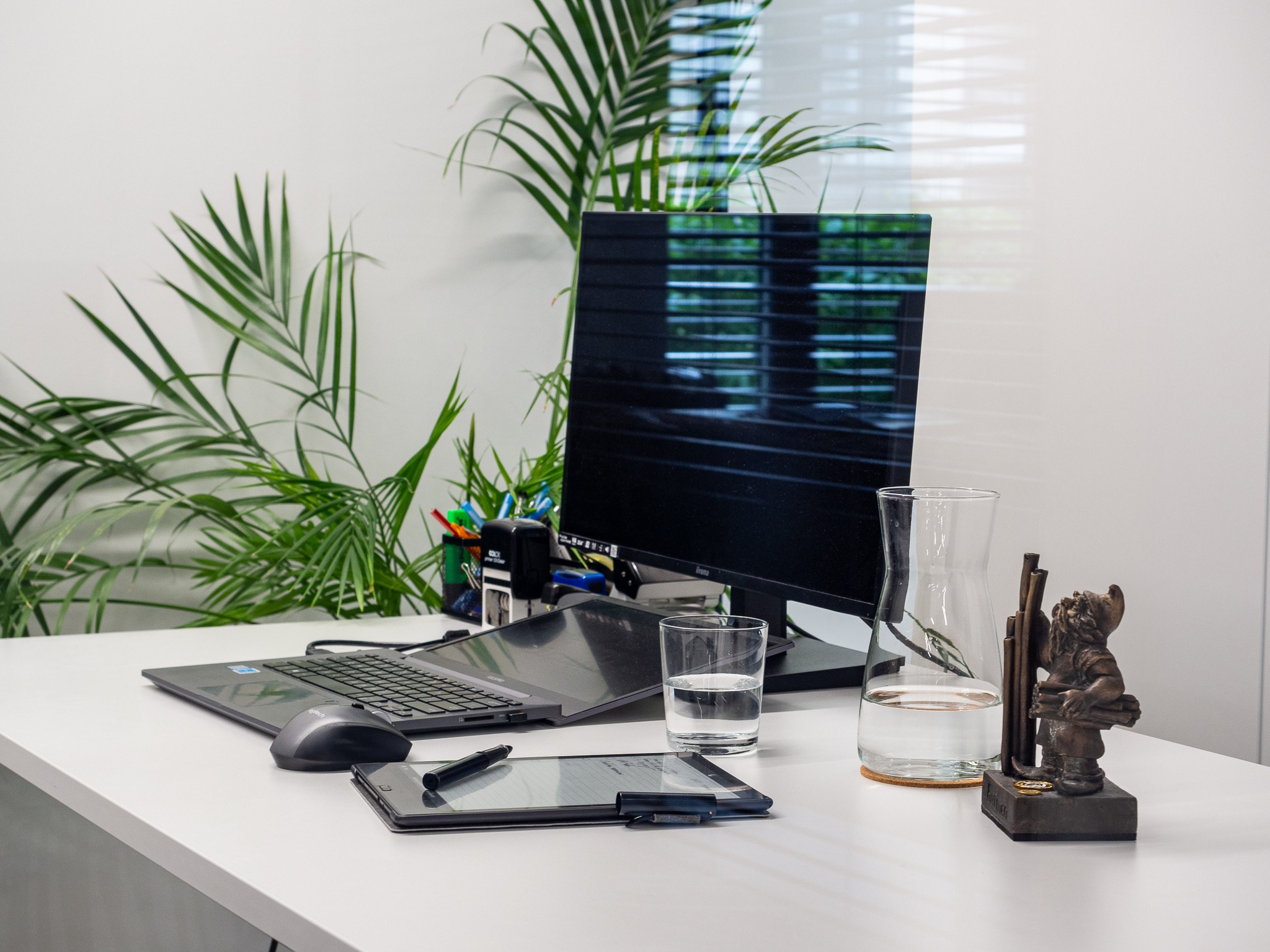 Nowoczesne biuro z laptopem, tabletem graficznym i szklanką wody na białym biurku, oświetlone naturalnym światłem, z zieloną rośliną w tle i statuetką.