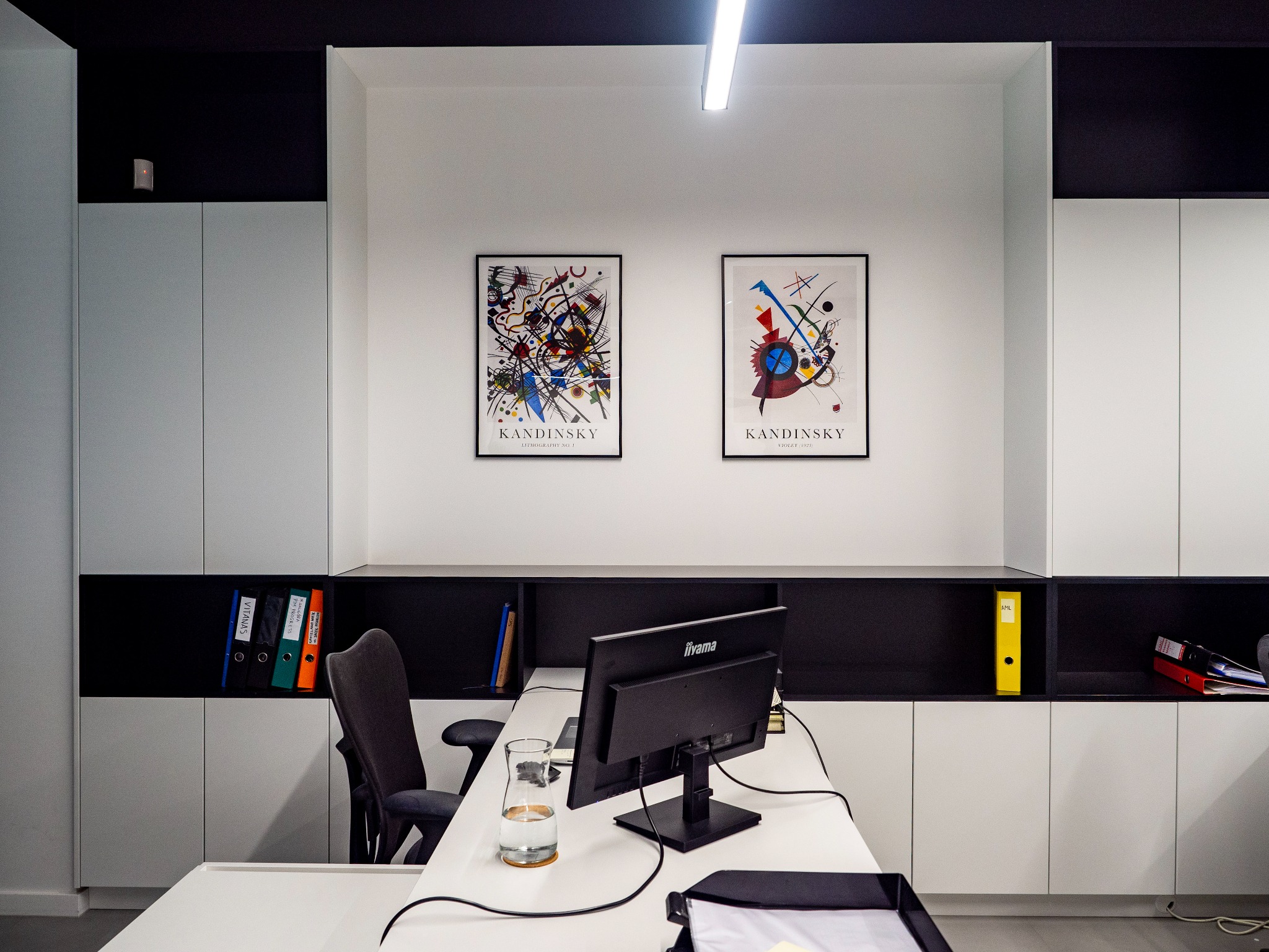 Nowoczesne biuro z białymi szafkami, czarnymi półkami, dwoma abstrakcyjnymi obrazami Kandinsky'ego na ścianie, biurkiem z monitorem Iiyama, szklanką wody i segregatorami w półkach.