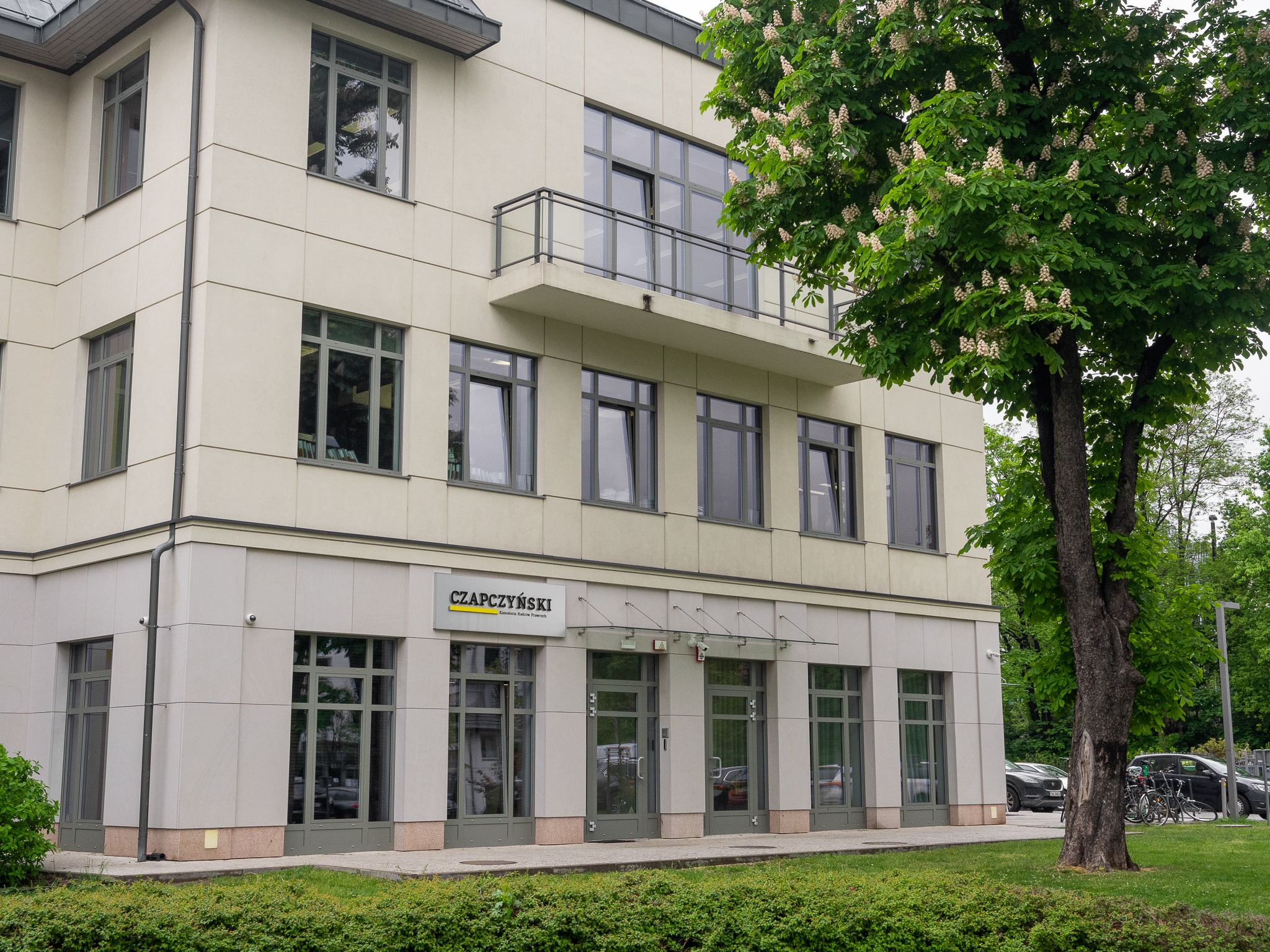 Elewacja budynku biurowego z szyldem kancelarii prawnej Czapczyński nad wejściem, widok z poziomu trawnika z kwitnącym kasztanowcem po prawej stronie