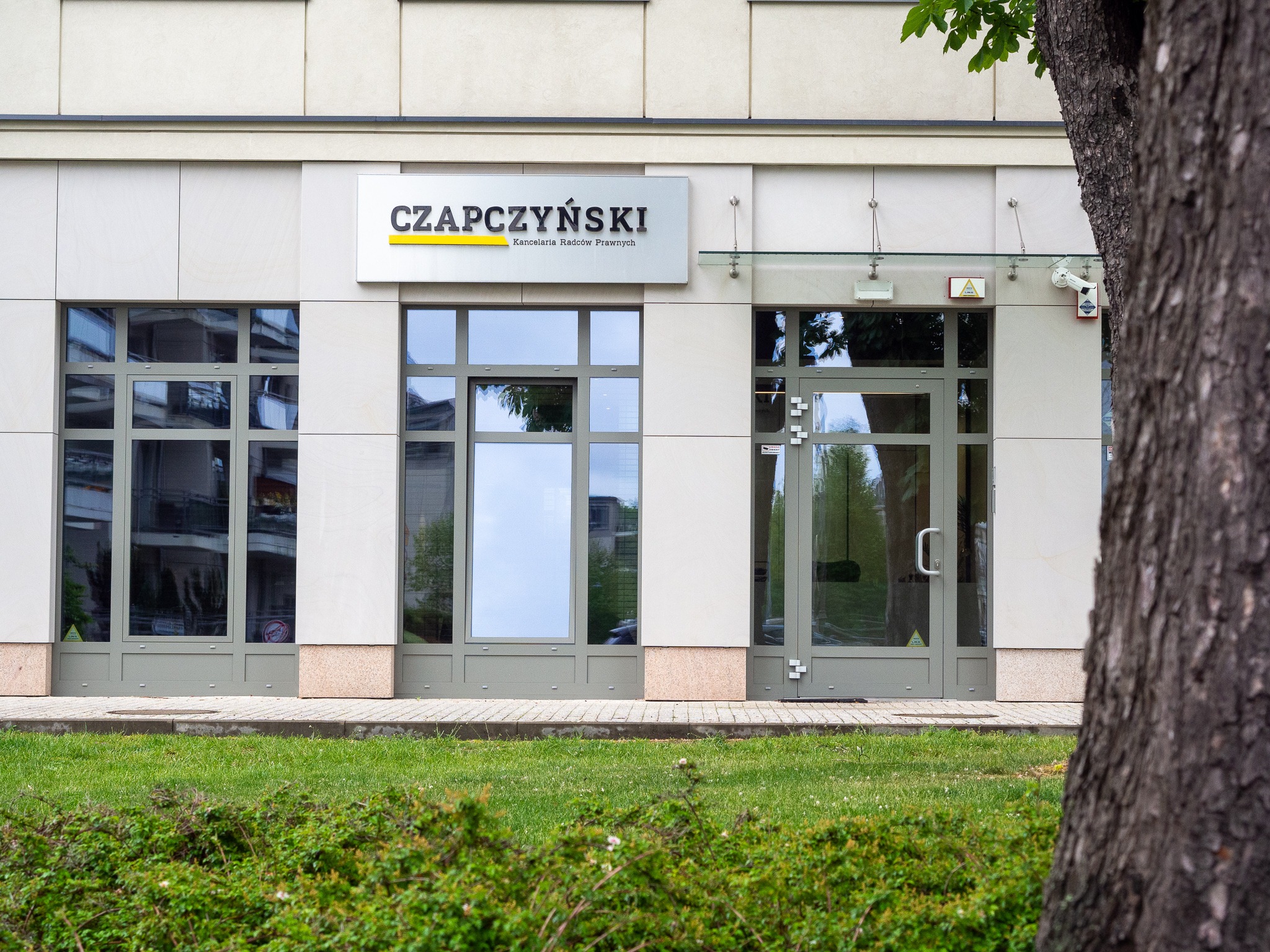 Fasada budynku z szyldem 'Czapczyński Kancelaria Radców Prawnych', widoczne okna z refleksami i szklane drzwi wejściowe z alarmem, fragment pnia drzewa po prawej stronie.