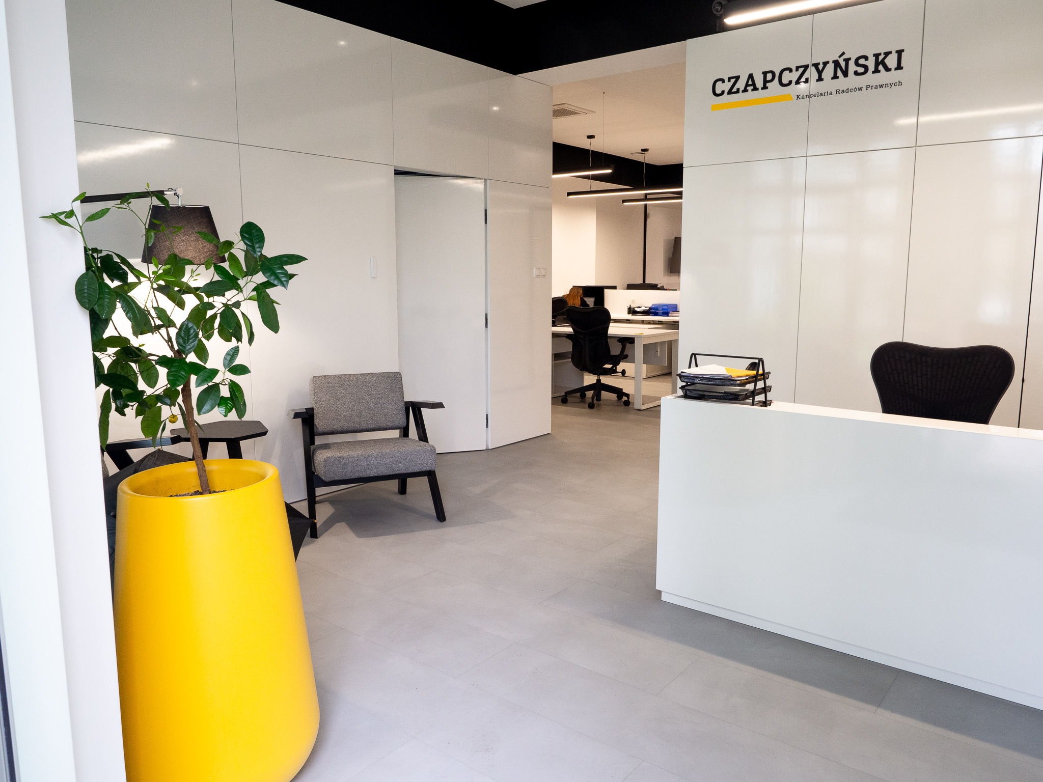 Nowoczesne wnętrze kancelarii prawnej z recepcją, szarym fotelem i dużą żółtą donicą z rośliną, widok na biura w tle.